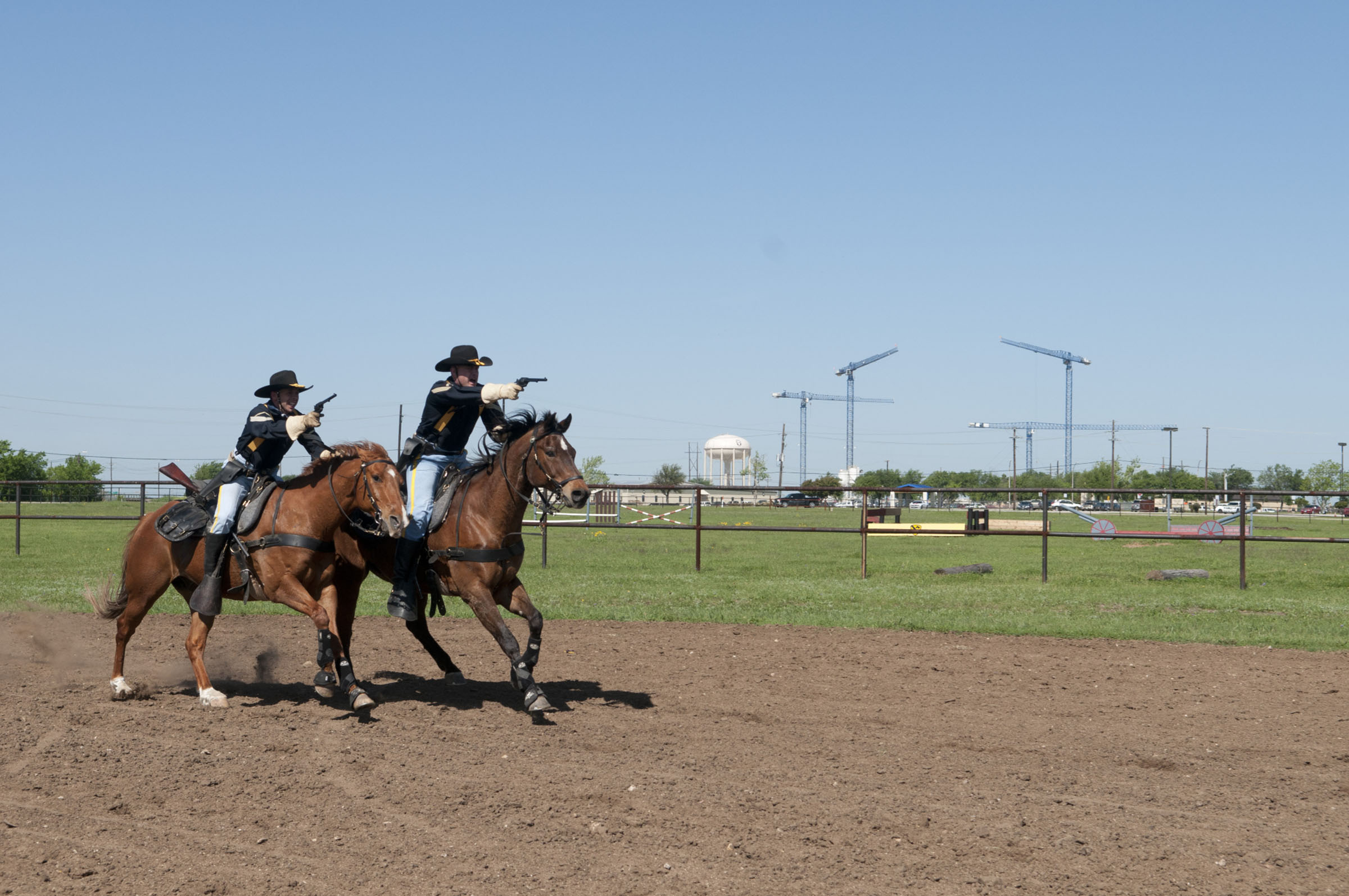 Two people ride on horseback on a dirt track while pointing revolvers in front of them.