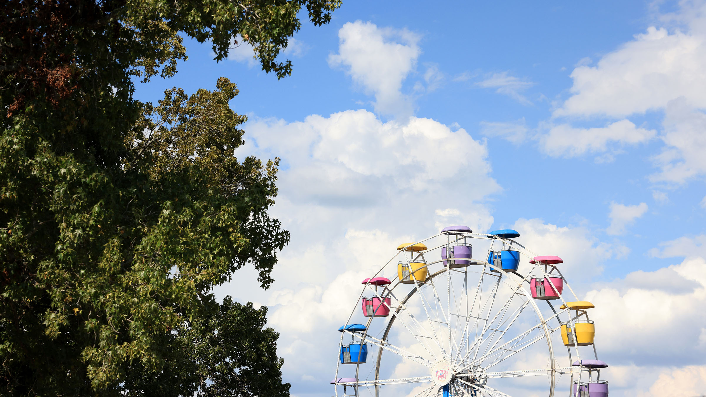 A ferris wheel is framed by green trees to the left, with a blue sky and clouds behind.
