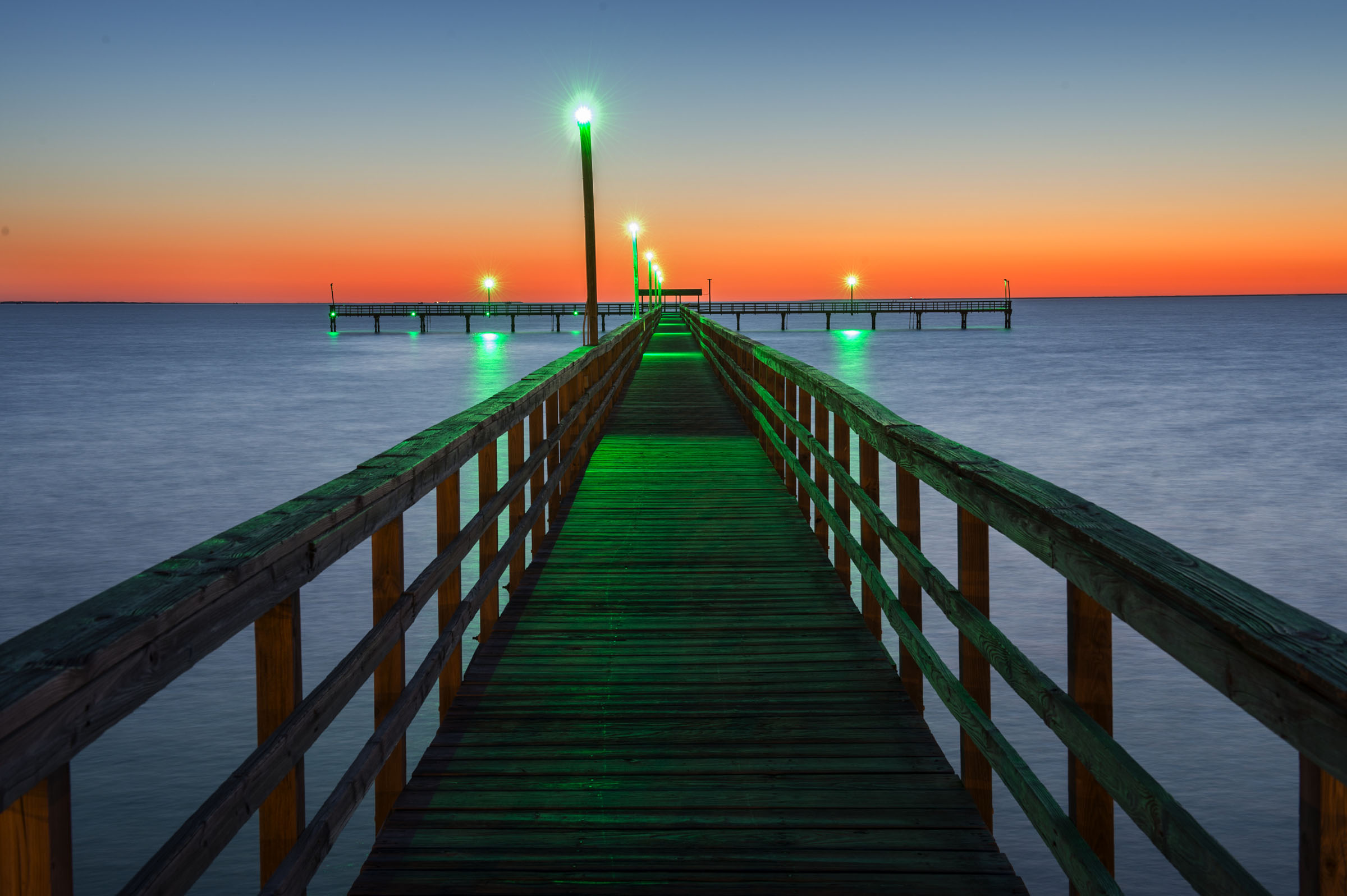 Green lights illuminate a pier leading to an orange sunset