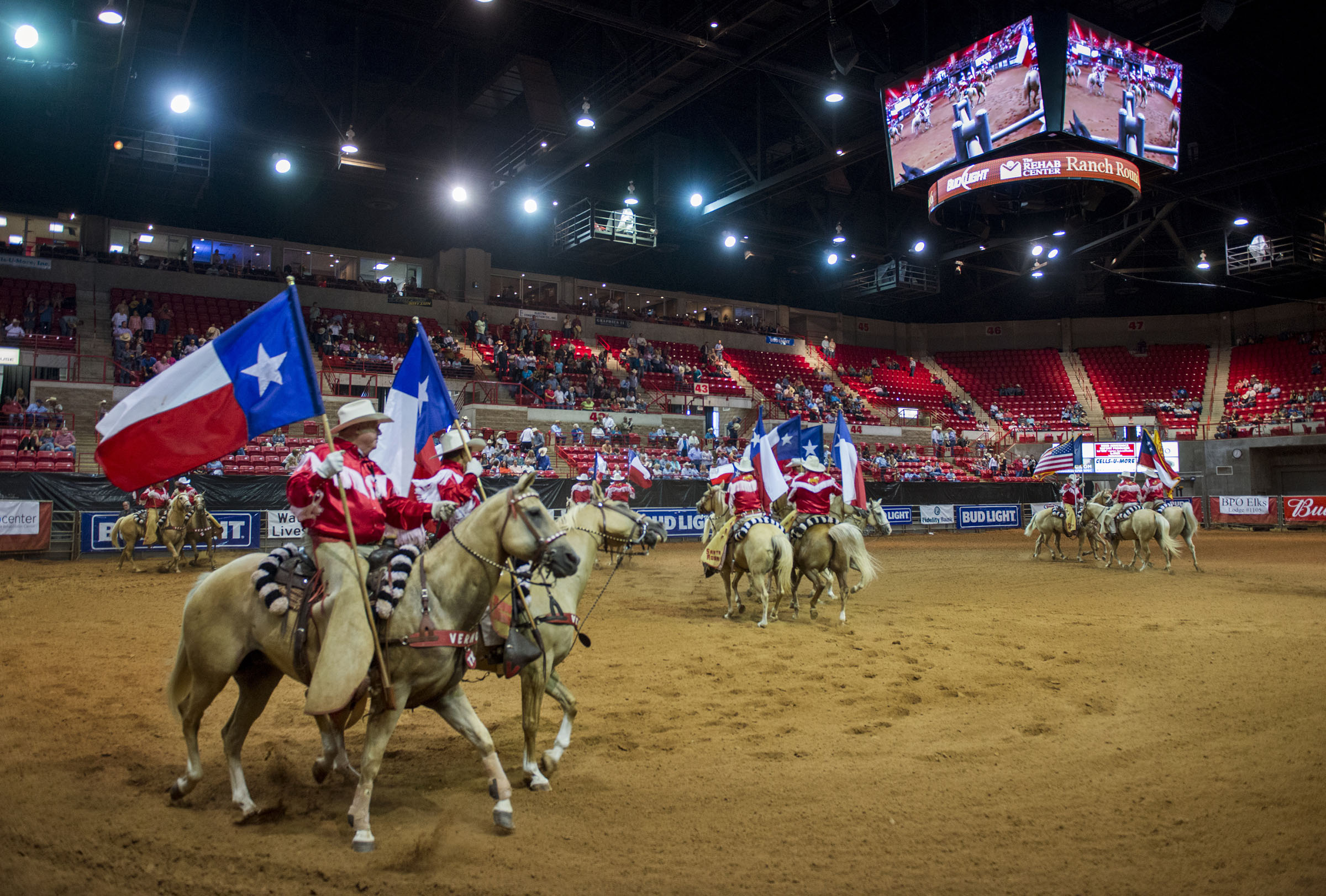 Riders on horseback carry Texas flags in a sports arena.