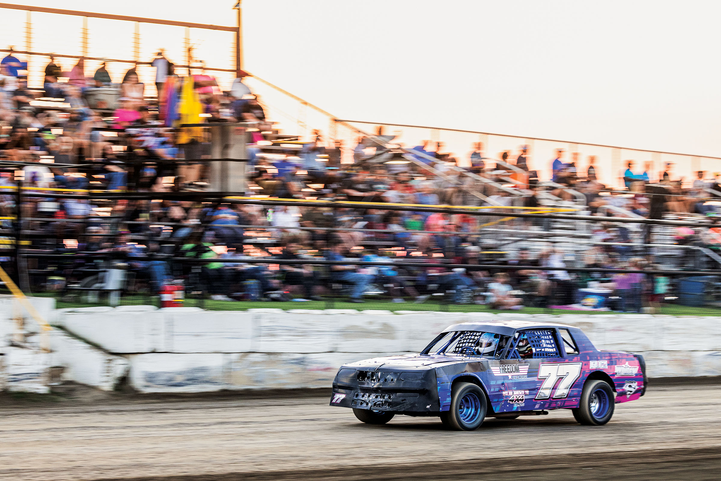 A purple stock car races down a racetrack in front of a crowd of spectators