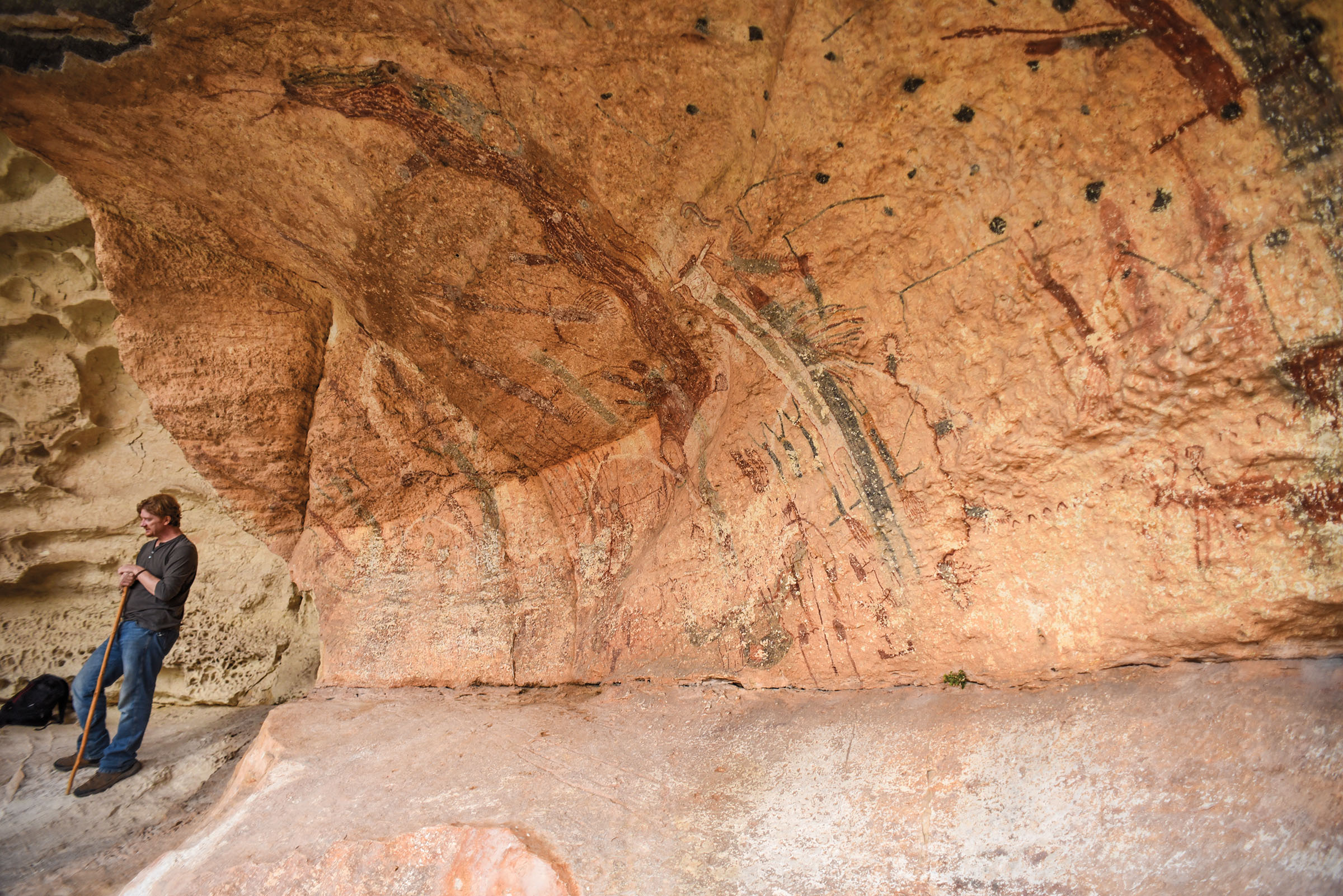 A guide stands next to a reddish rock wall with numerous cave drawings