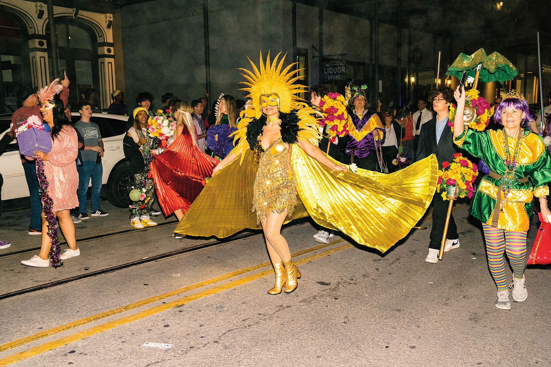 A woman wearing a long gold cape and gold crown walks down a street in a parade