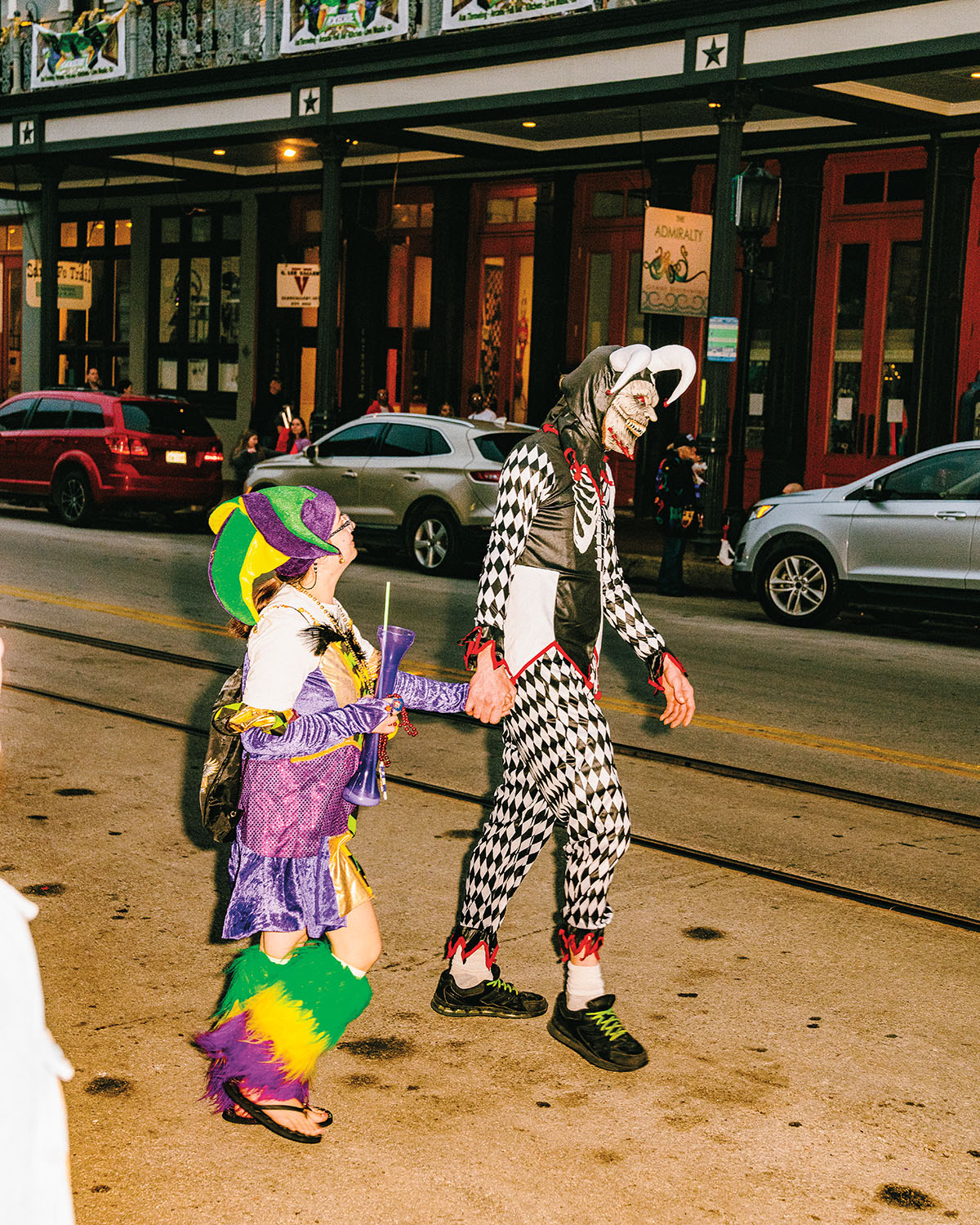 A person in a purple, green, and yellow jester costume walks next to a taller person in a black and white costume with a mask