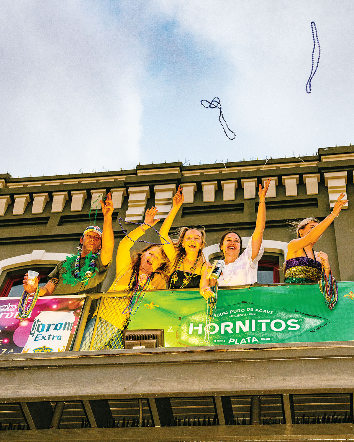 A group of people throw beads from a balcony, decorated and lit in bright yellow and green color