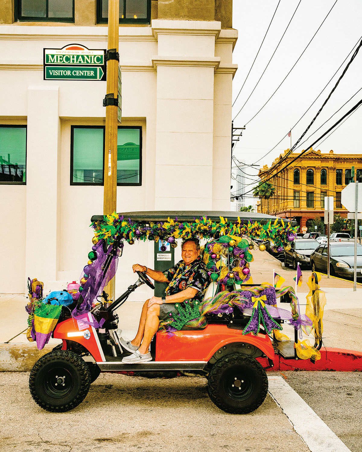 A man drives a golf cart decorated with green, purple, and gold decorations on a city street