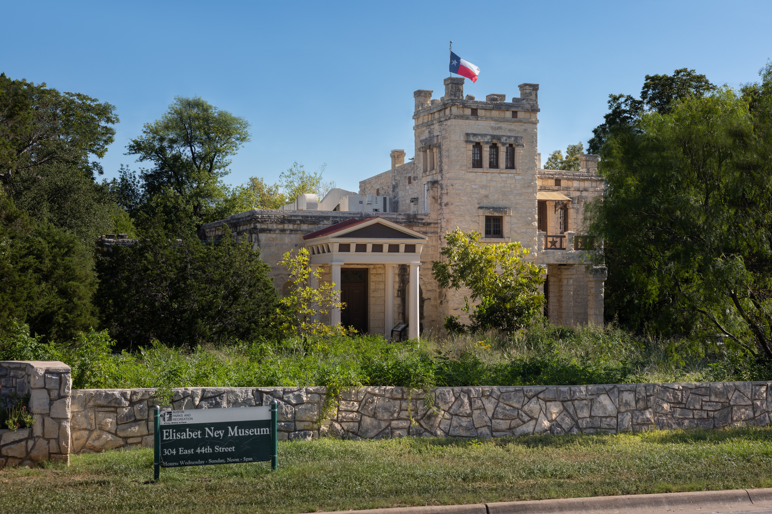 Exterior shot of the three-story stone museum with sign and stone wall in the foreground and Texas flag on flagpole waving at top of tower. 