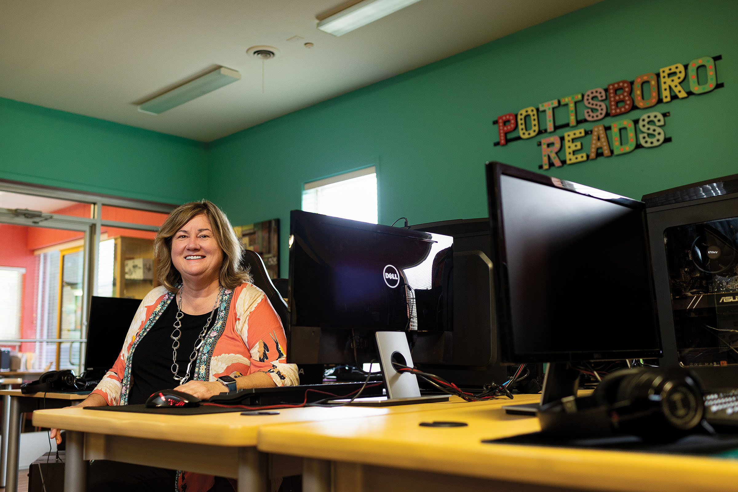 A woman sits in front of a computer in a room with numerous computers and lettering on the wall reading "Pottsboro Reads"