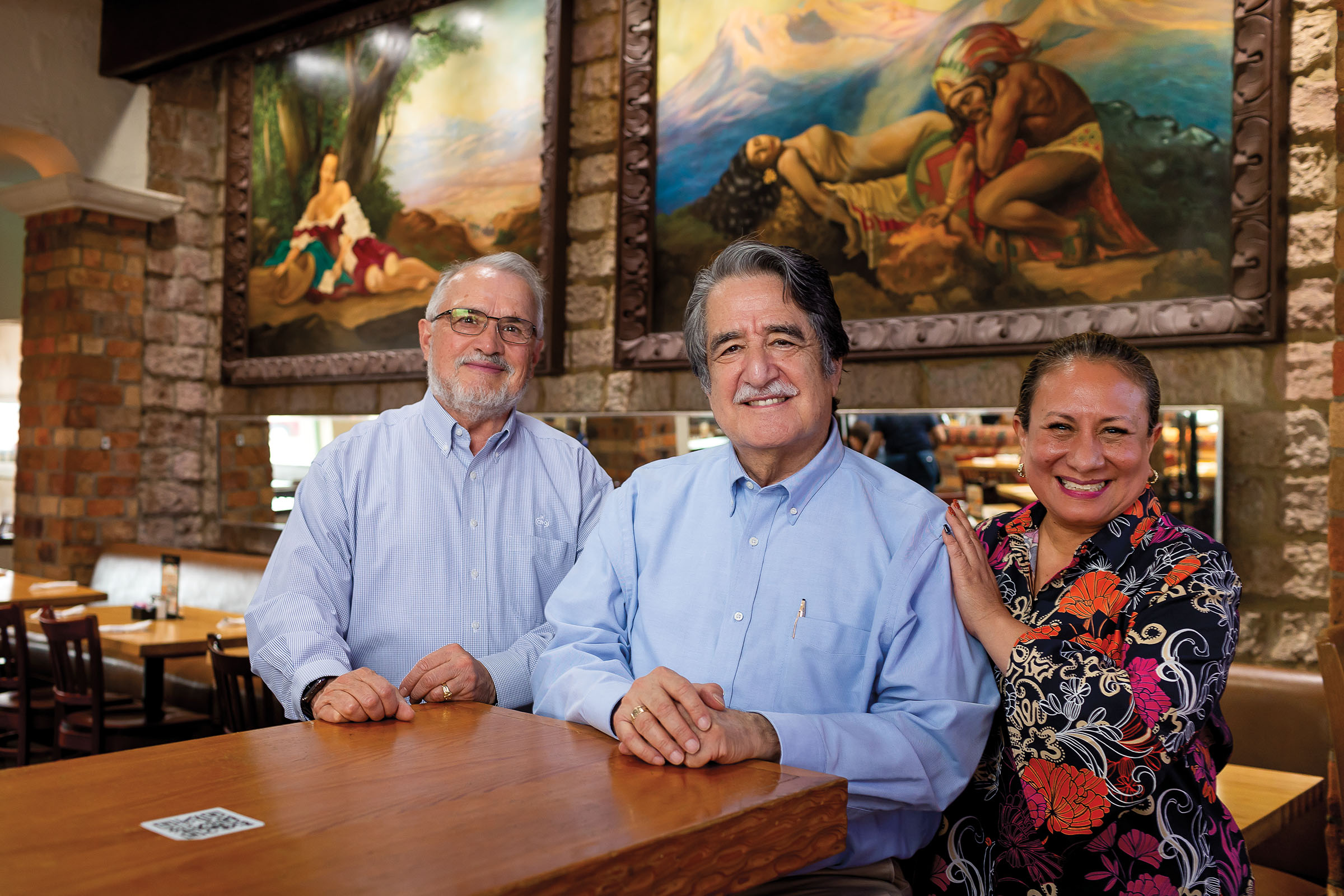 A group of three people pose, standing at a wooden table top in front of large artwork on the wall behind