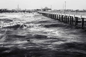 A black and white photo of large waves crashing against a long pier