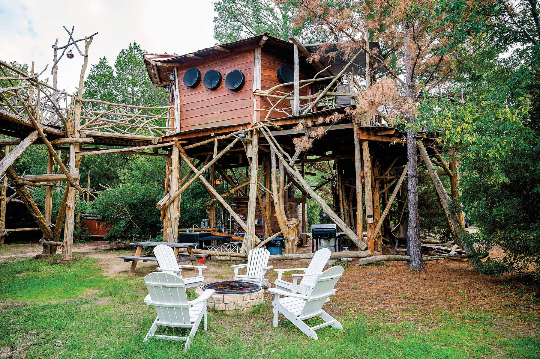 A treehouse built on tall wooden stilts, with white Adirondack chairs in the front 