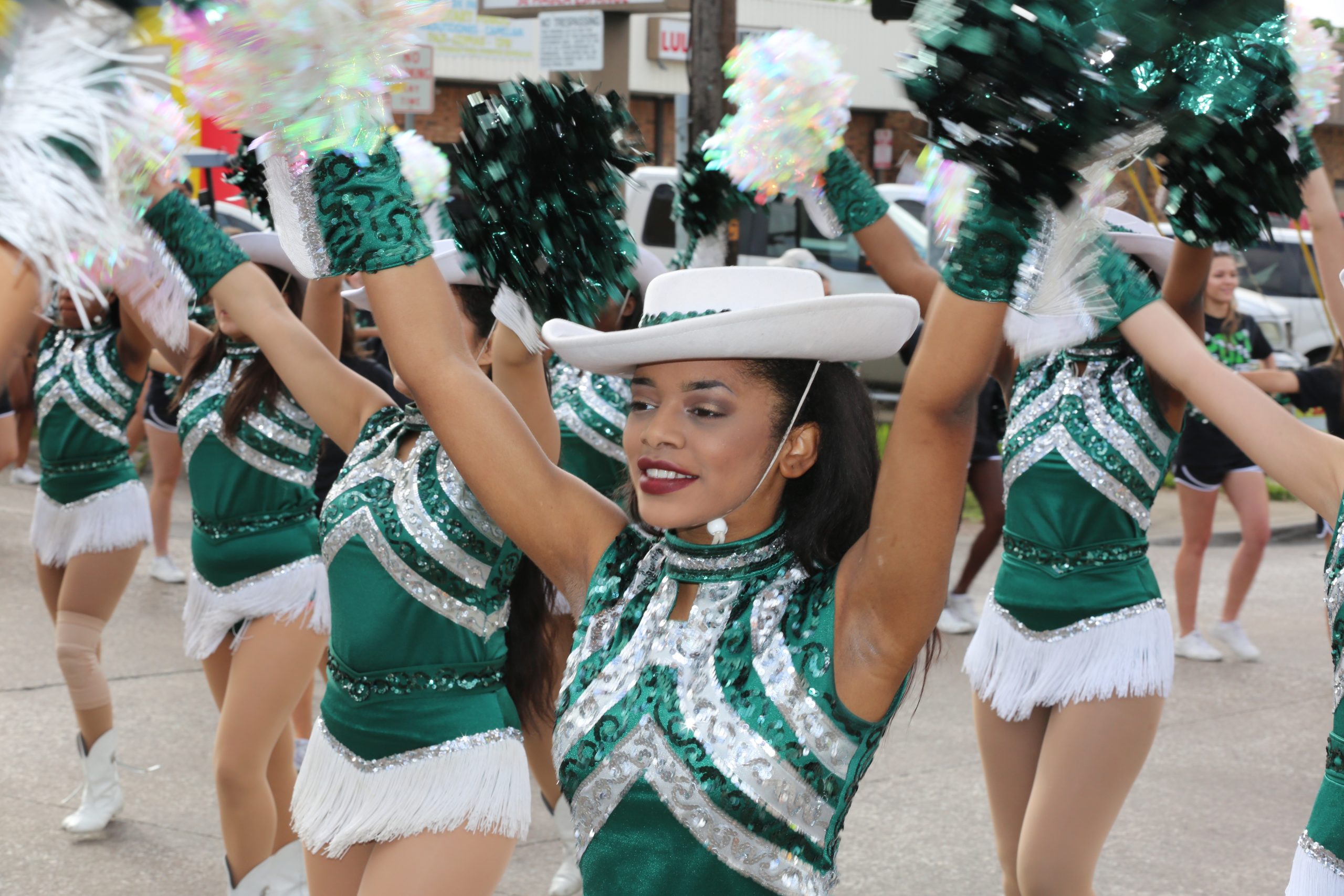With other drill team members in background, young woman wearing a white cowboy hat and green and white sequined costume holds up pompoms while in the parade. 