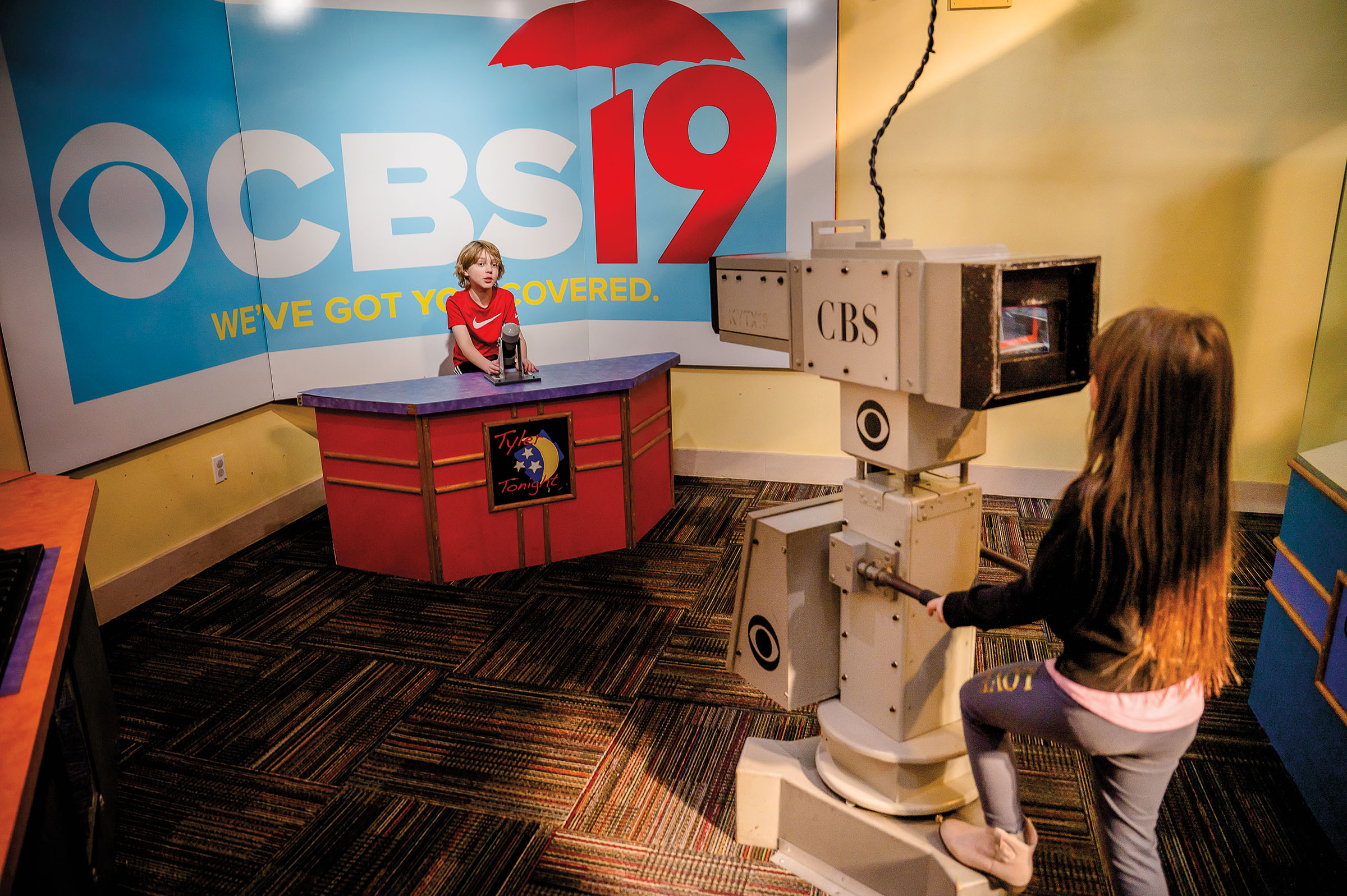 Two young people use a replica TV set including a large news camera, desk, and background reading 