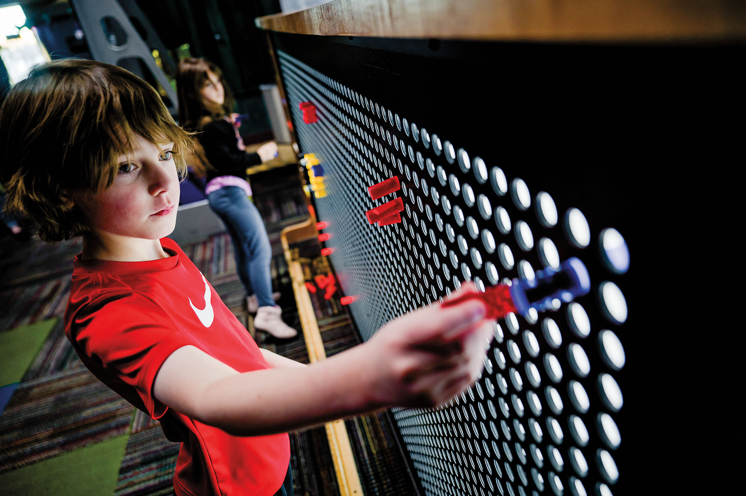 A young person in a red shirt attaches bright plastic pegs to a light-up wall