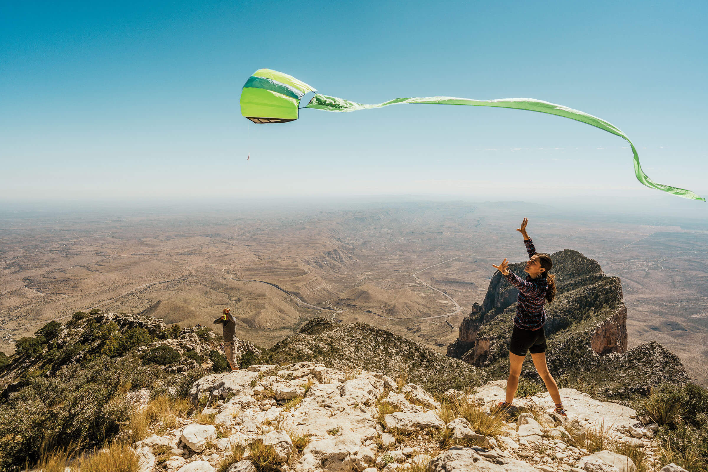 A person flies a green kite over a mountain peak on a blue sky day