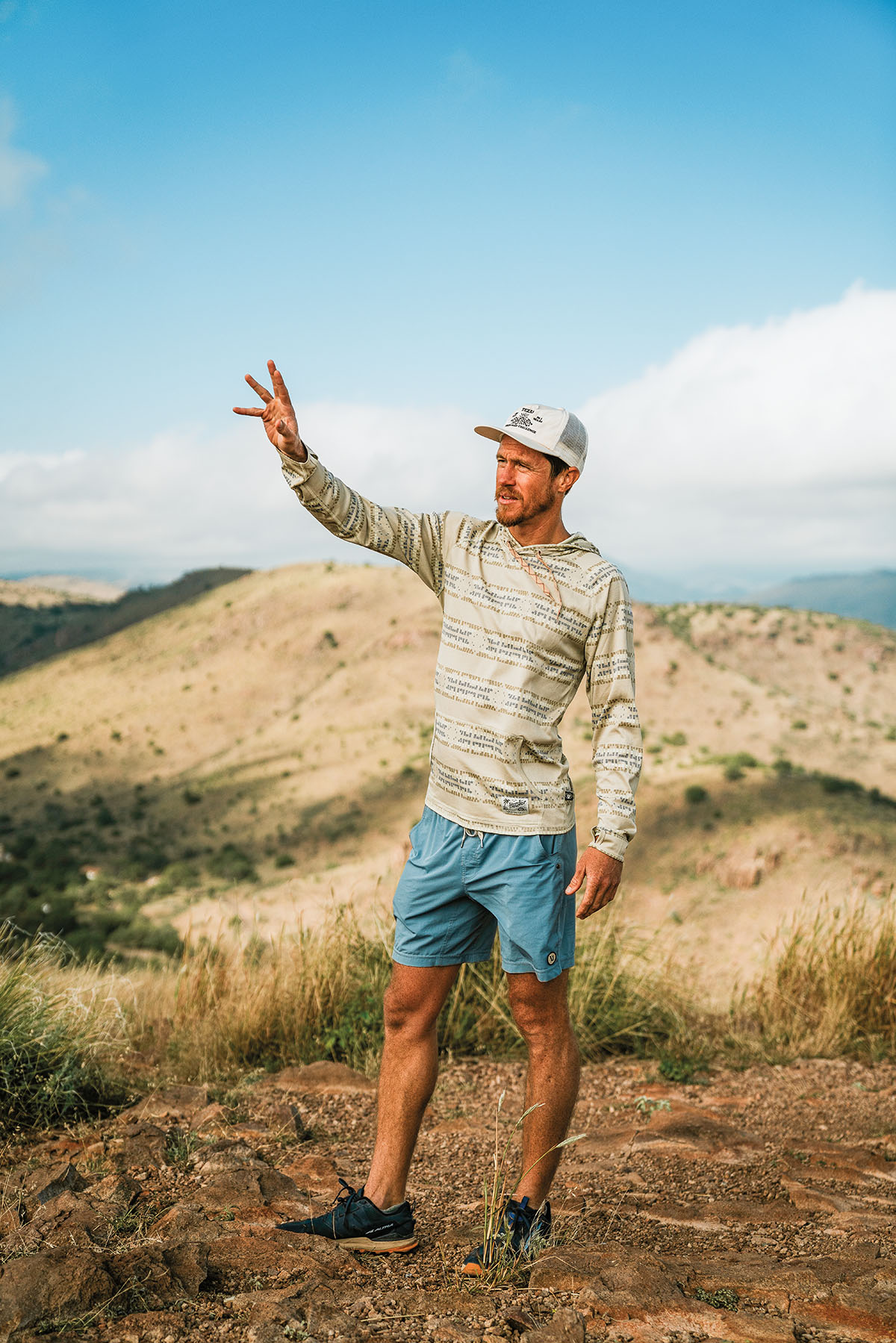 A man wearing a baseball cap and shorts stands in a desert mountain environment