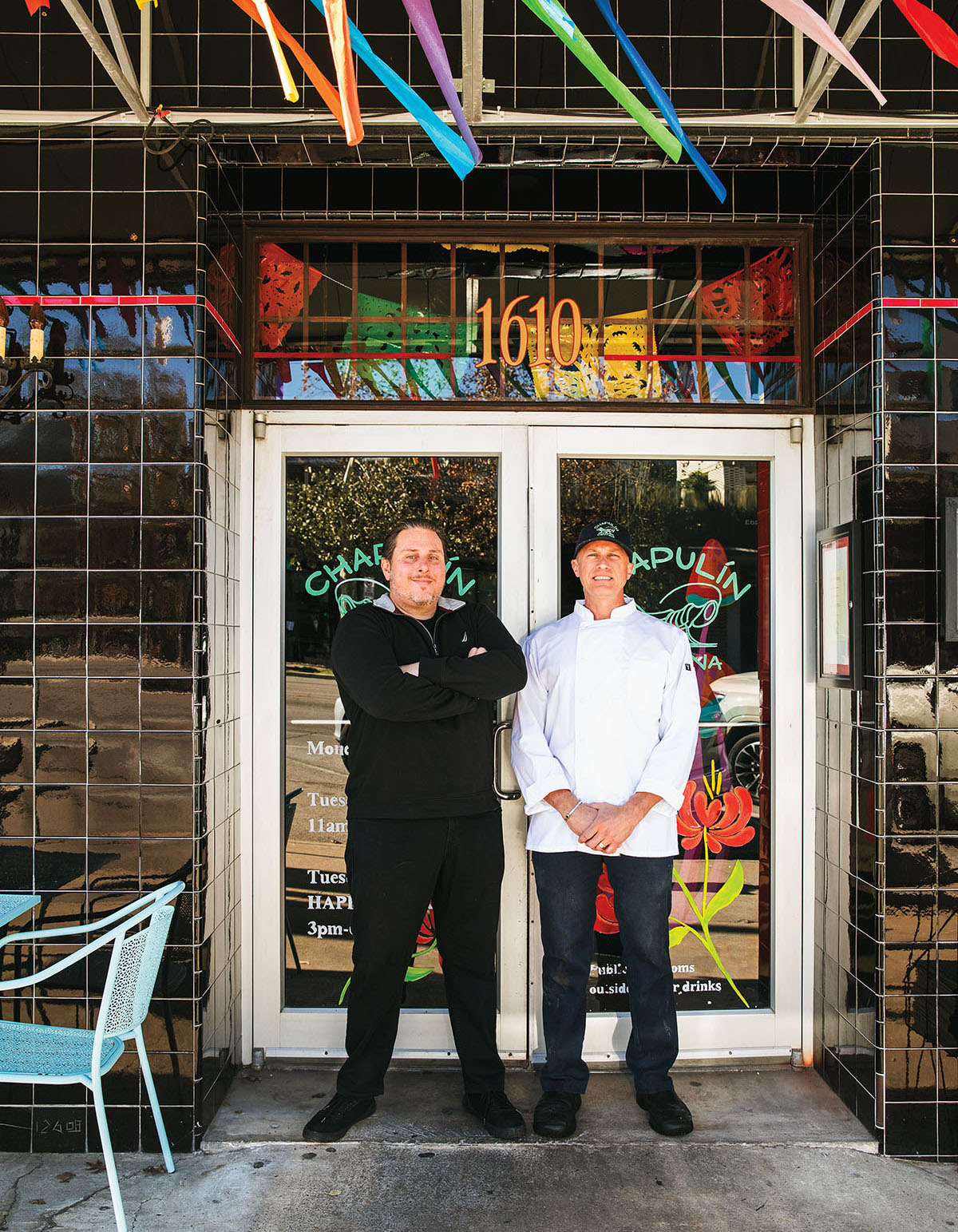 Two men in chef's coats stand out front of the glass doors of a restaurant