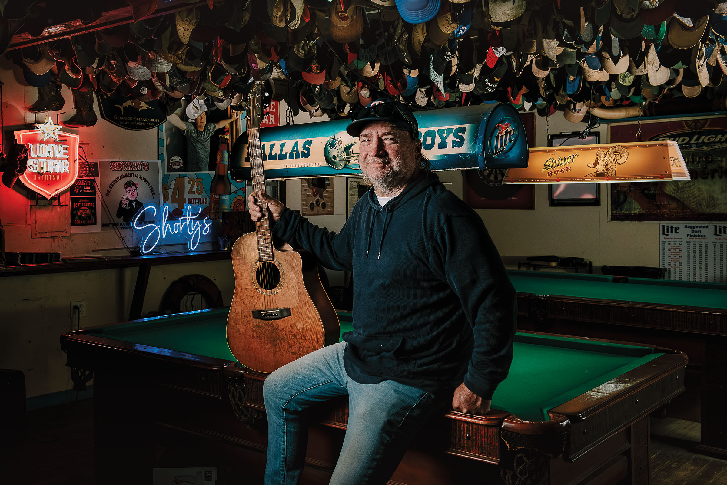 A man with a guitar leans on a pool table in a dimly-lit bar