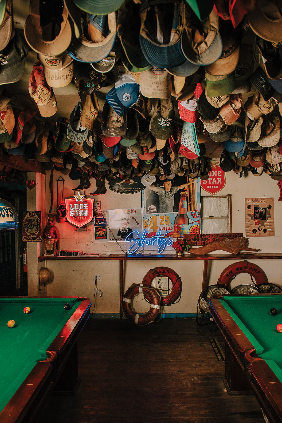 A large collection of hats pack the ceiling of a bar above velvet green pool tables