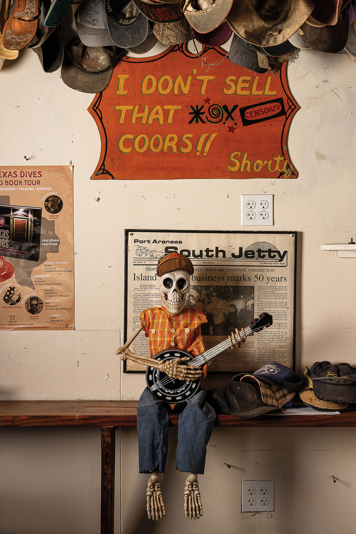 A skeleton wearing a shirt and holding a guitar in front of a collection of posters and memorabilia including a sign reading 