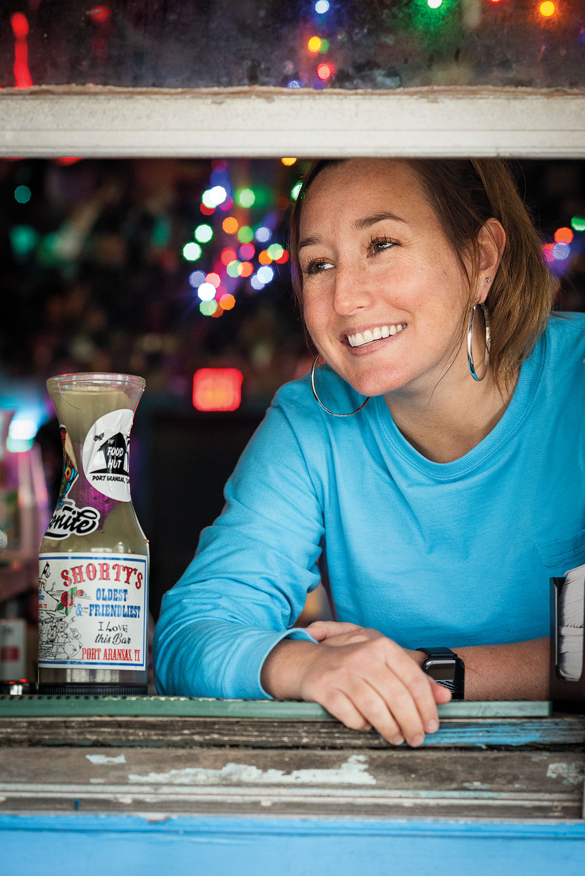 A woman wearing a blue shirt smiles next to a bar tip jar