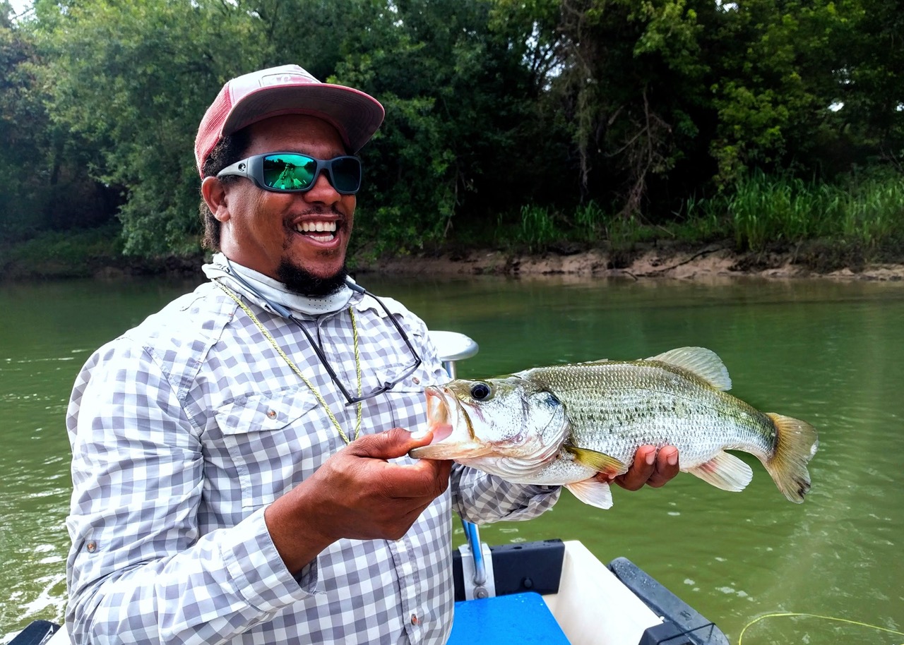 Alvin Dedeaux, wearing a cap, shades, and plaid shirt, holds up a large fish while smiling. Background has water and trees.