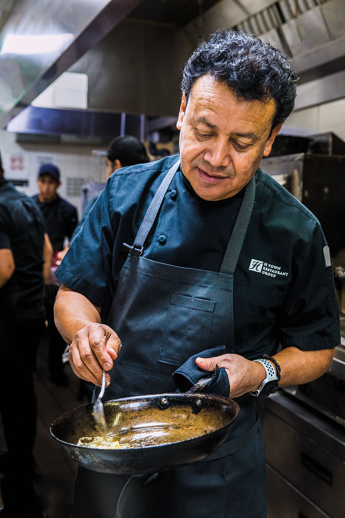A chef wearing a dark blue apron mixes a sauce in a pan