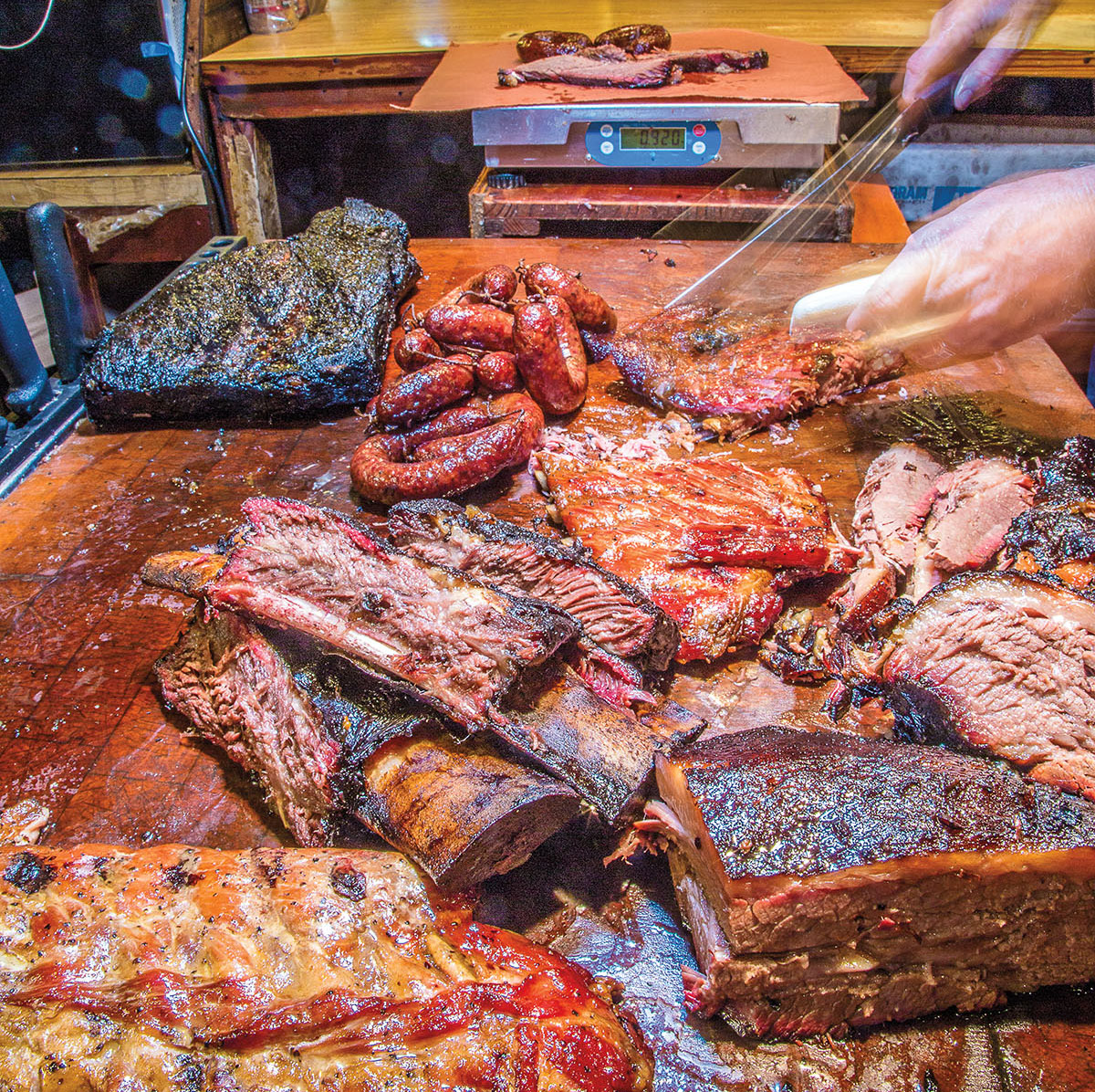 A person slices meat around numerous barbecue offerings in front of a small scale