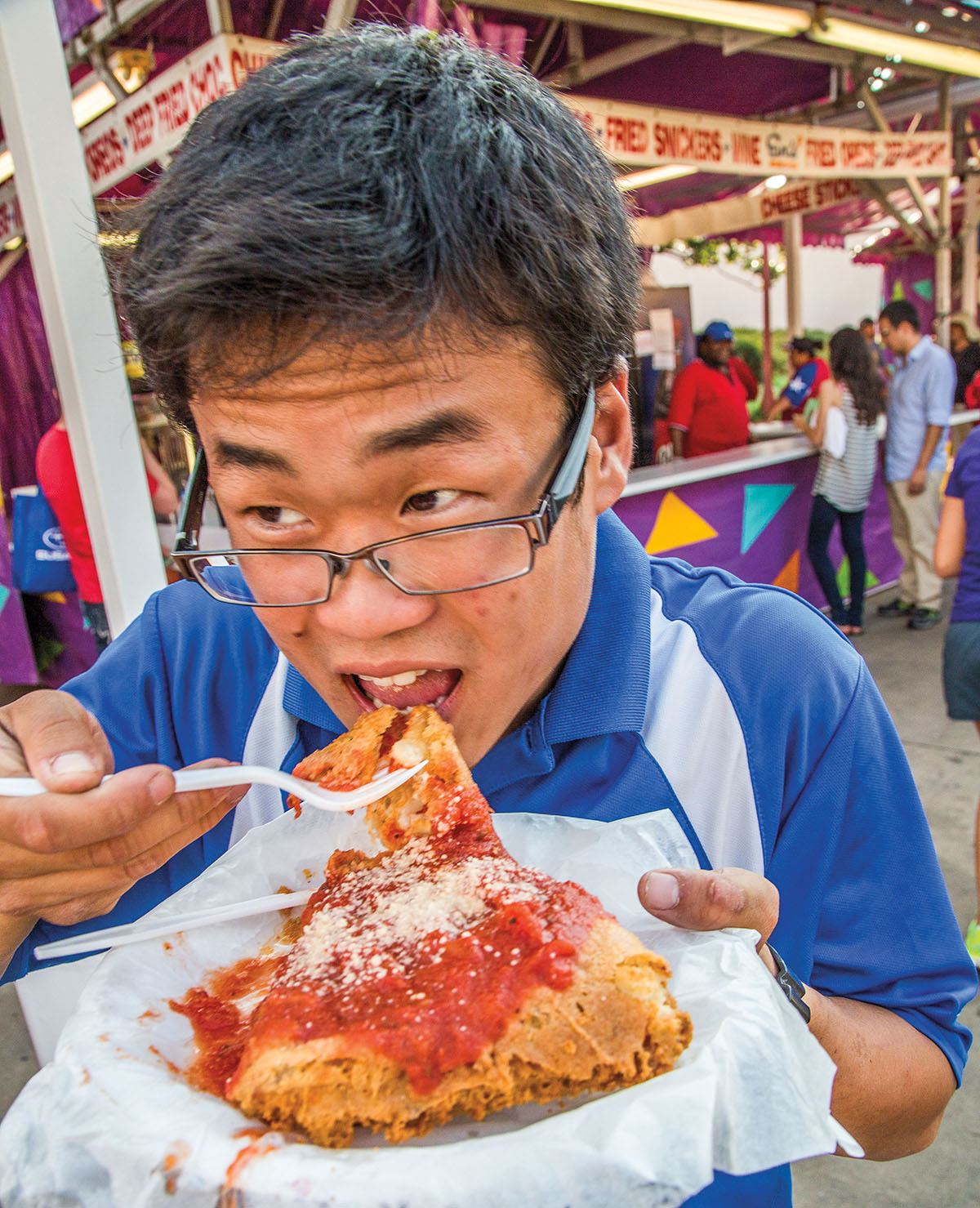 A person in a blue shirt eats a large fried item at a fair