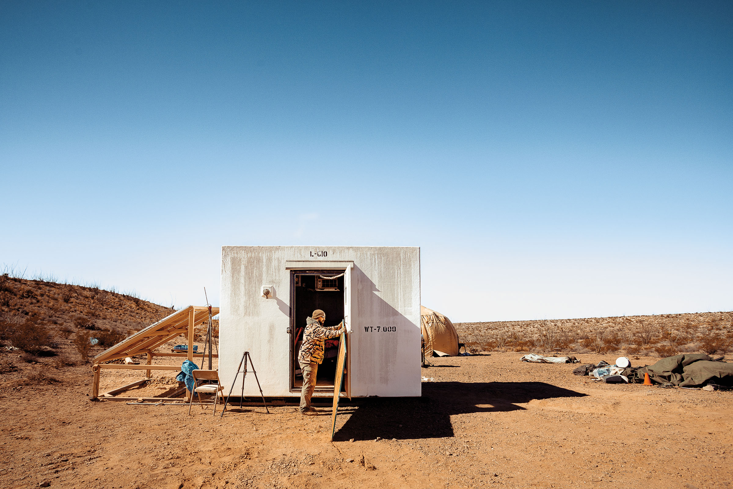 A man holds open the door of a small white structure in a desert landscape under blue sky