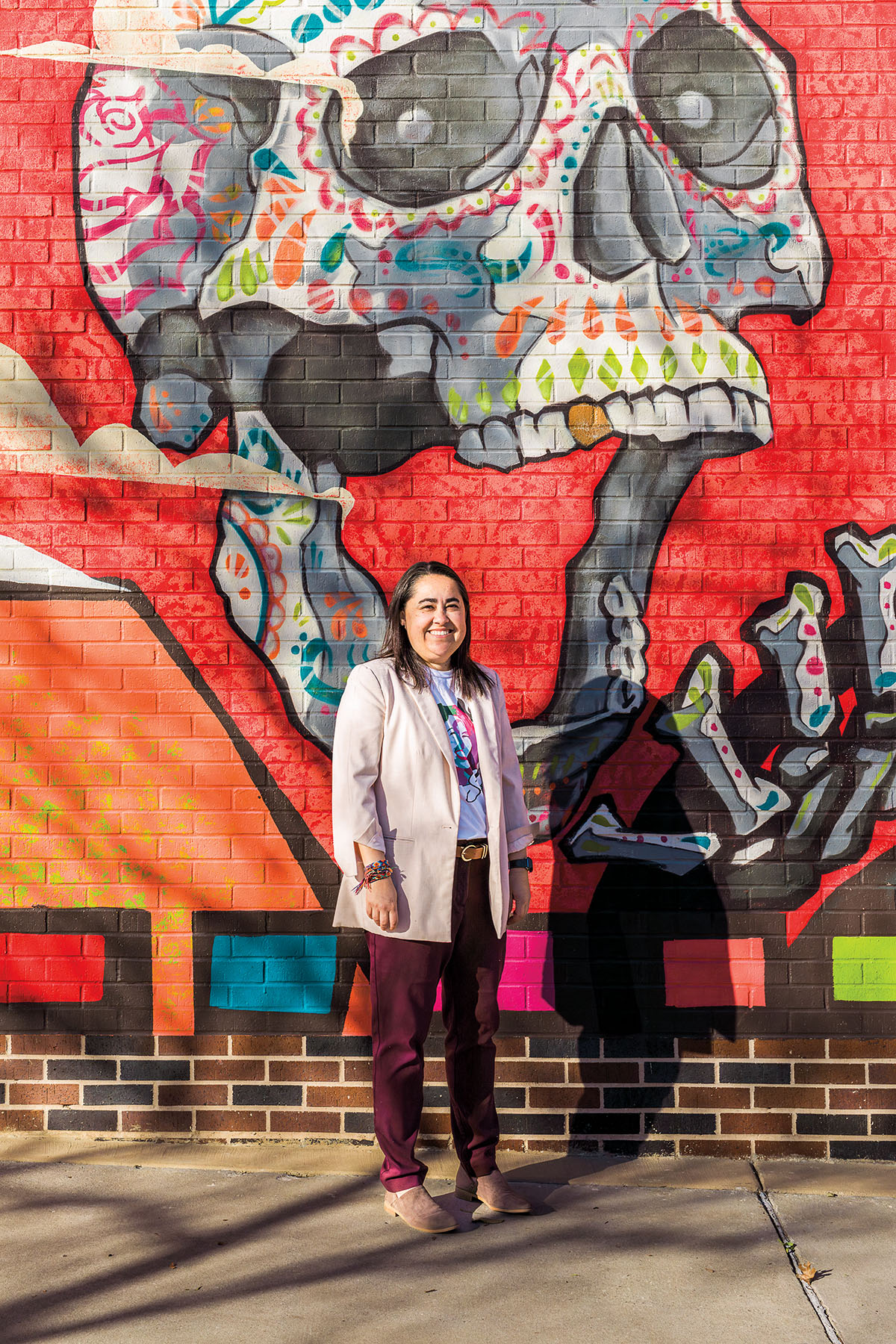 A woman wearing a light pink blazer stands in front of a brightly painted mural of a skull