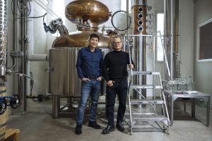 Two men stand in front of large steel brewing equipment