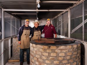 Three men stand inside an enclosed space behind a brick pit