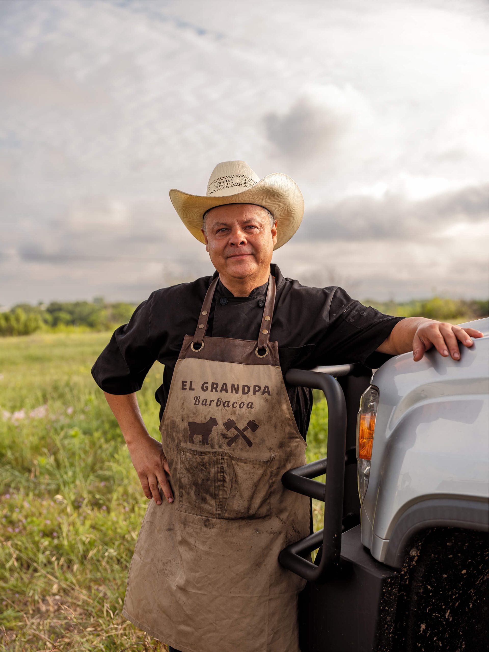 A man in a cowboy hat and apron stands in a field beside a truck