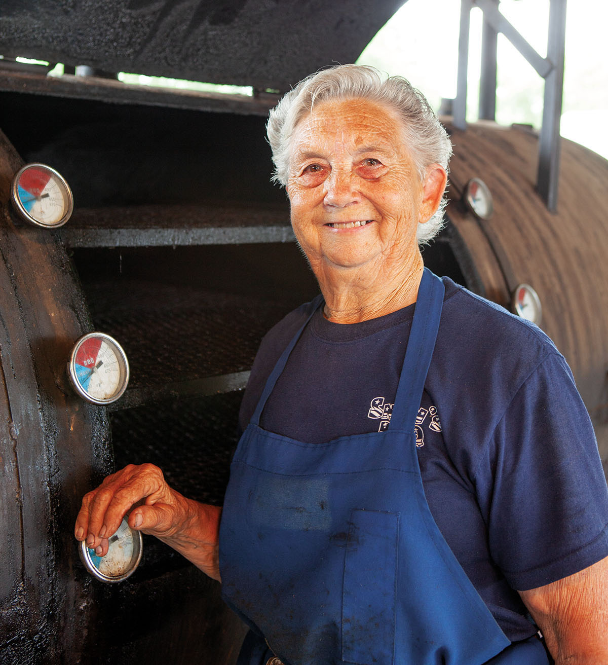 A woman wearing a blue apron stands in front of a large barbecue pit