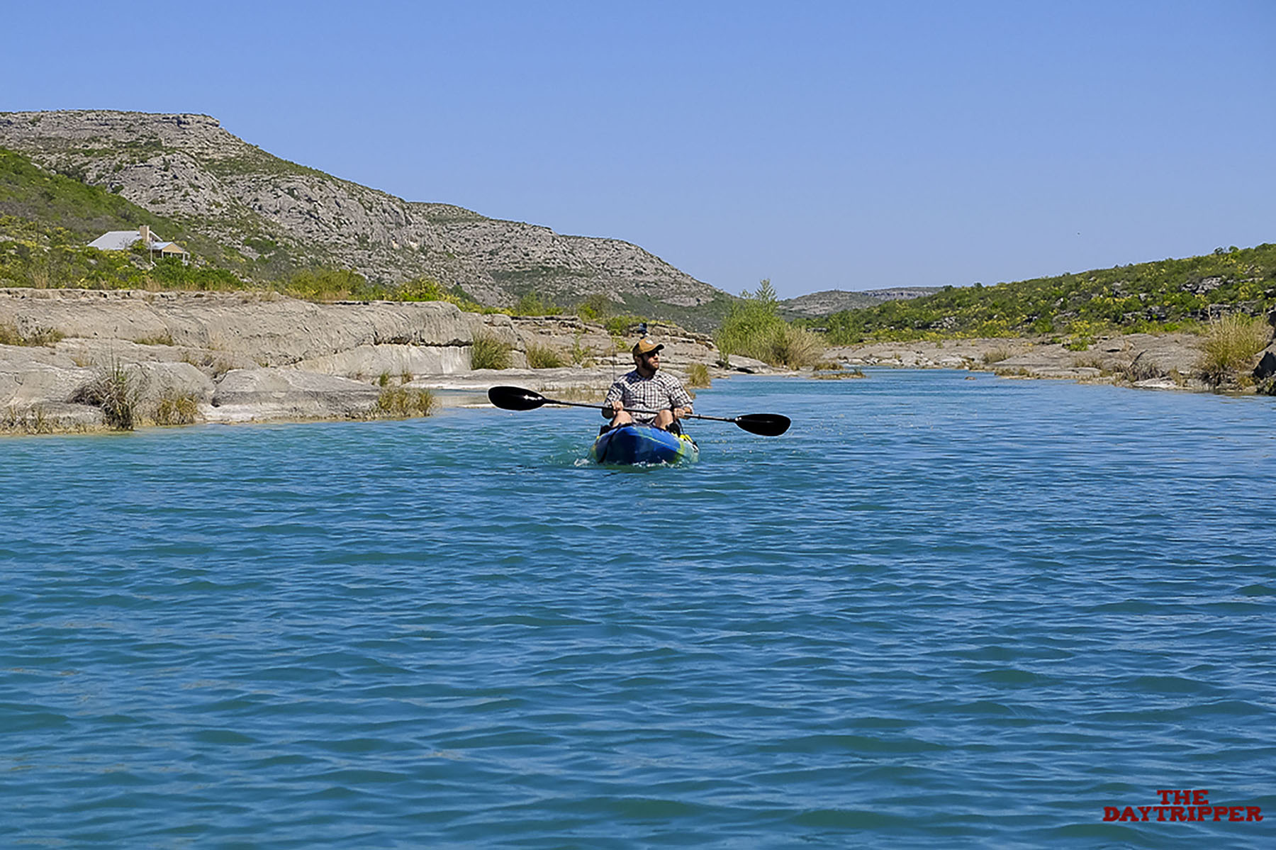 A man paddles a kayak down bright blue water with a hilly desert landscape in the background