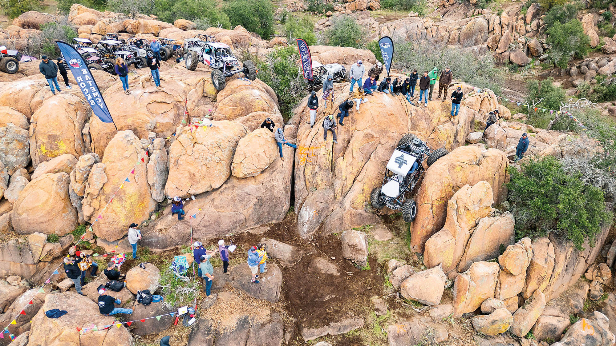 A group of people stand on tan rocks looking at a 4x4 vehicle attempting to scale the rock 