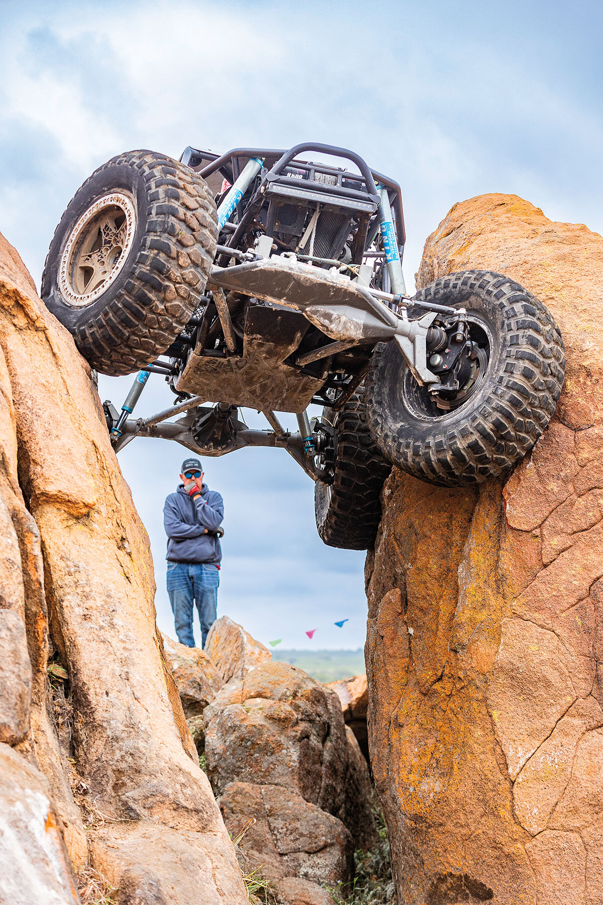 A person stands between two vertical rocks as a large 4x4 vehicle looms overhead