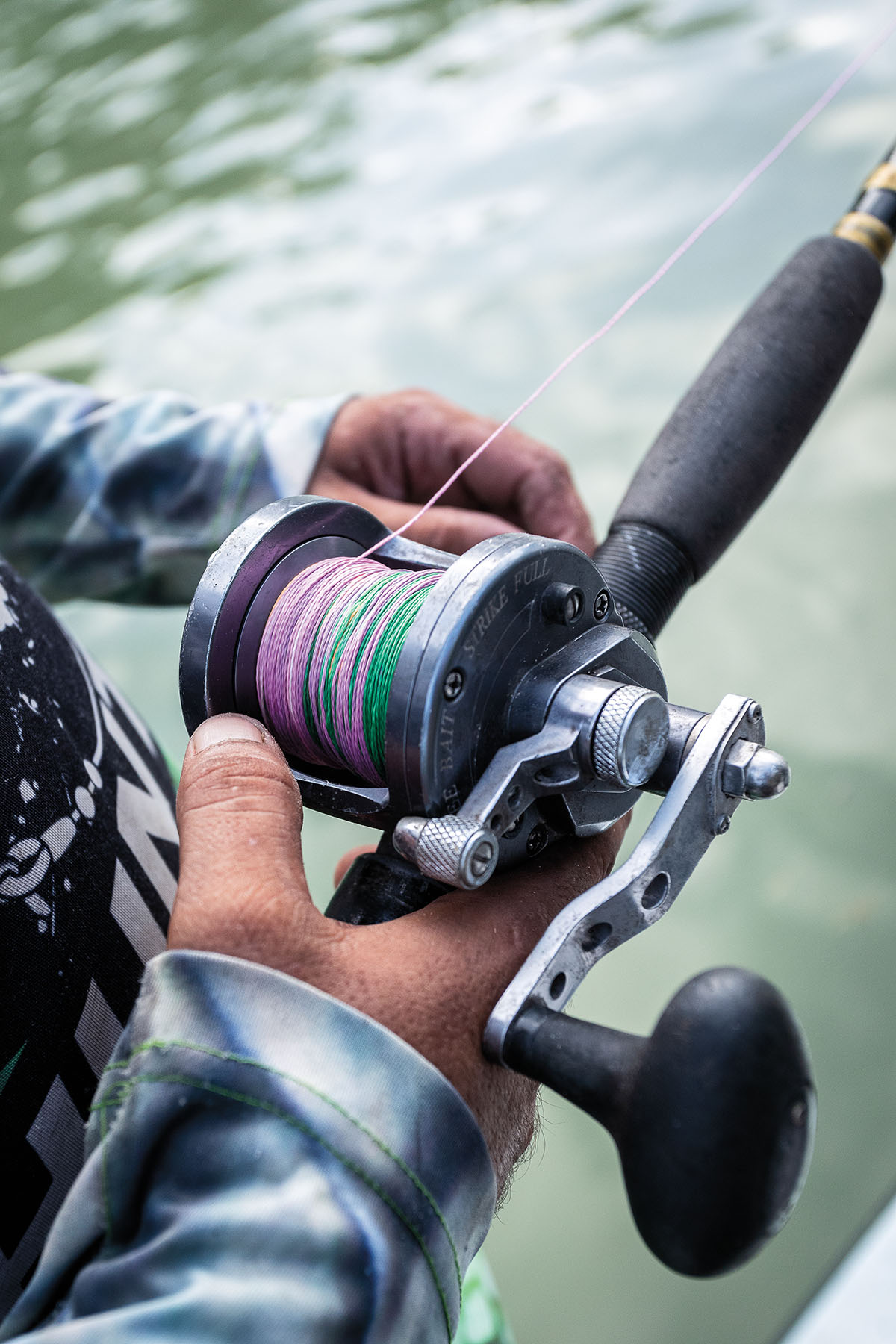 A person's hands hold a fishing reel with bring pink line above the water
