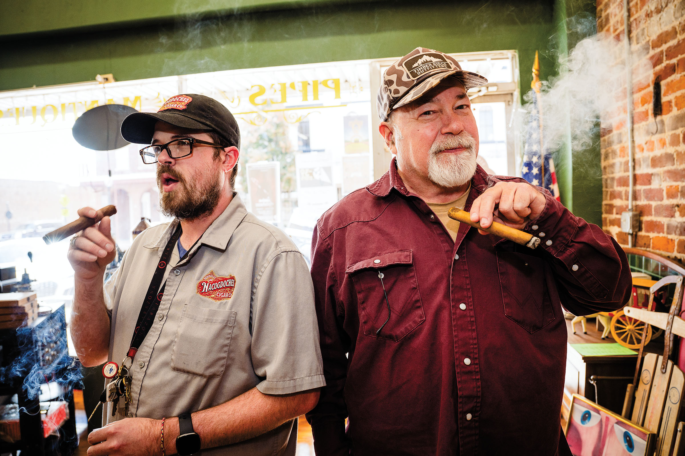 Two men stand next to each other with cigars inside of a brightly-lit store