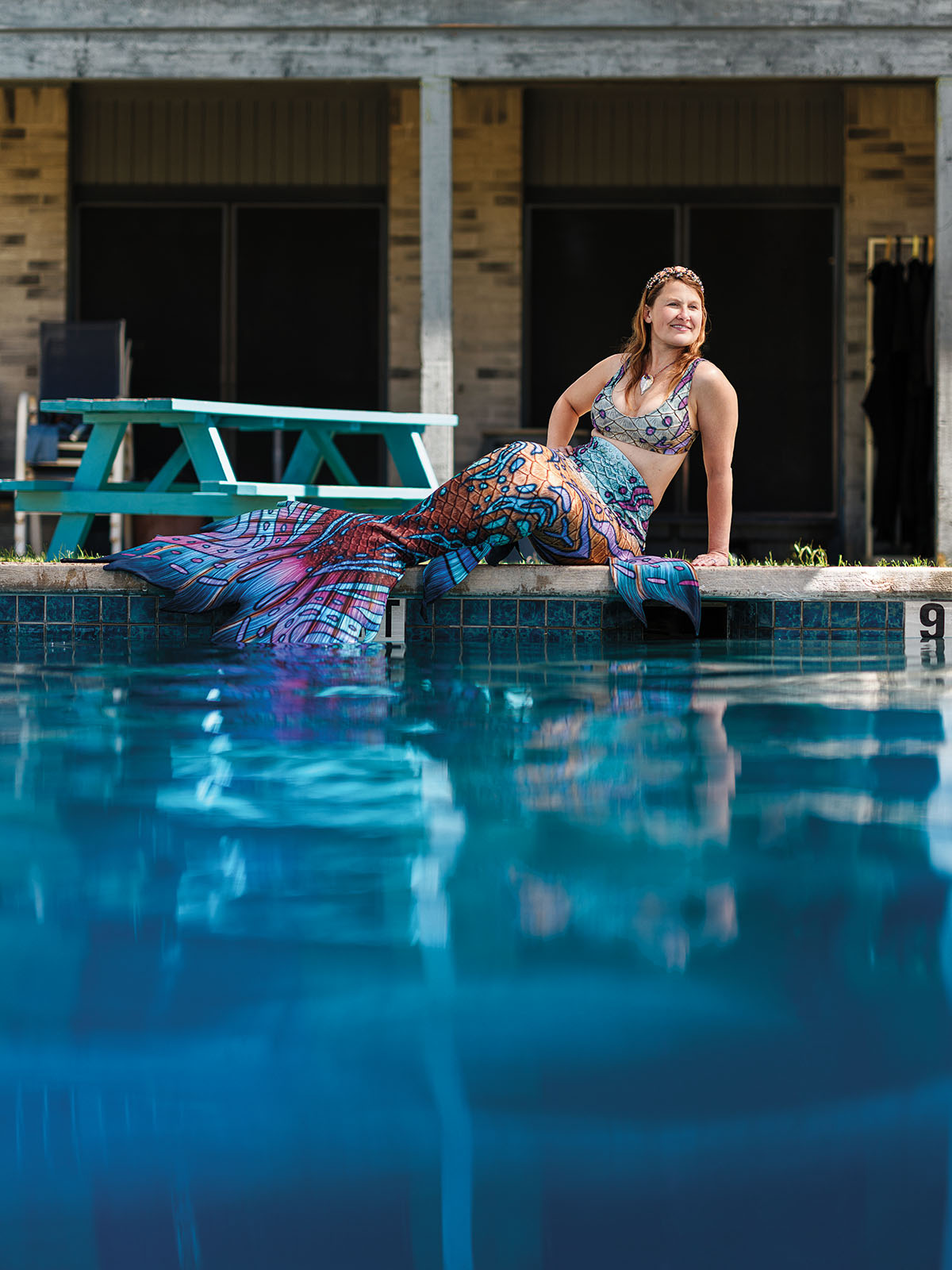A woman wearing a mermaid costume with a large, colorful tail sits by the side of a bright blue pool