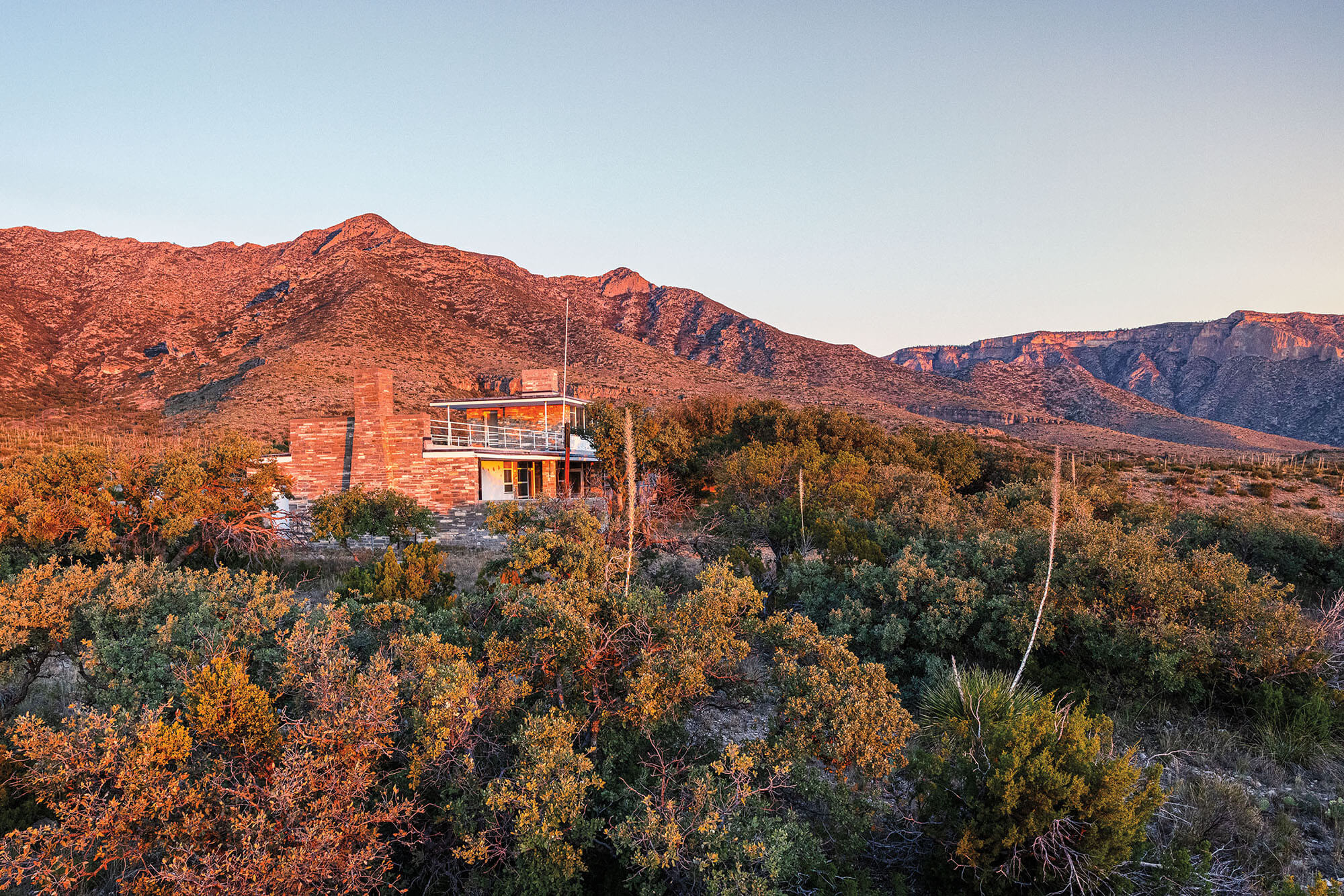 A large brick, midcentury home built in a desert landscape in front of a large rust-colored mountain
