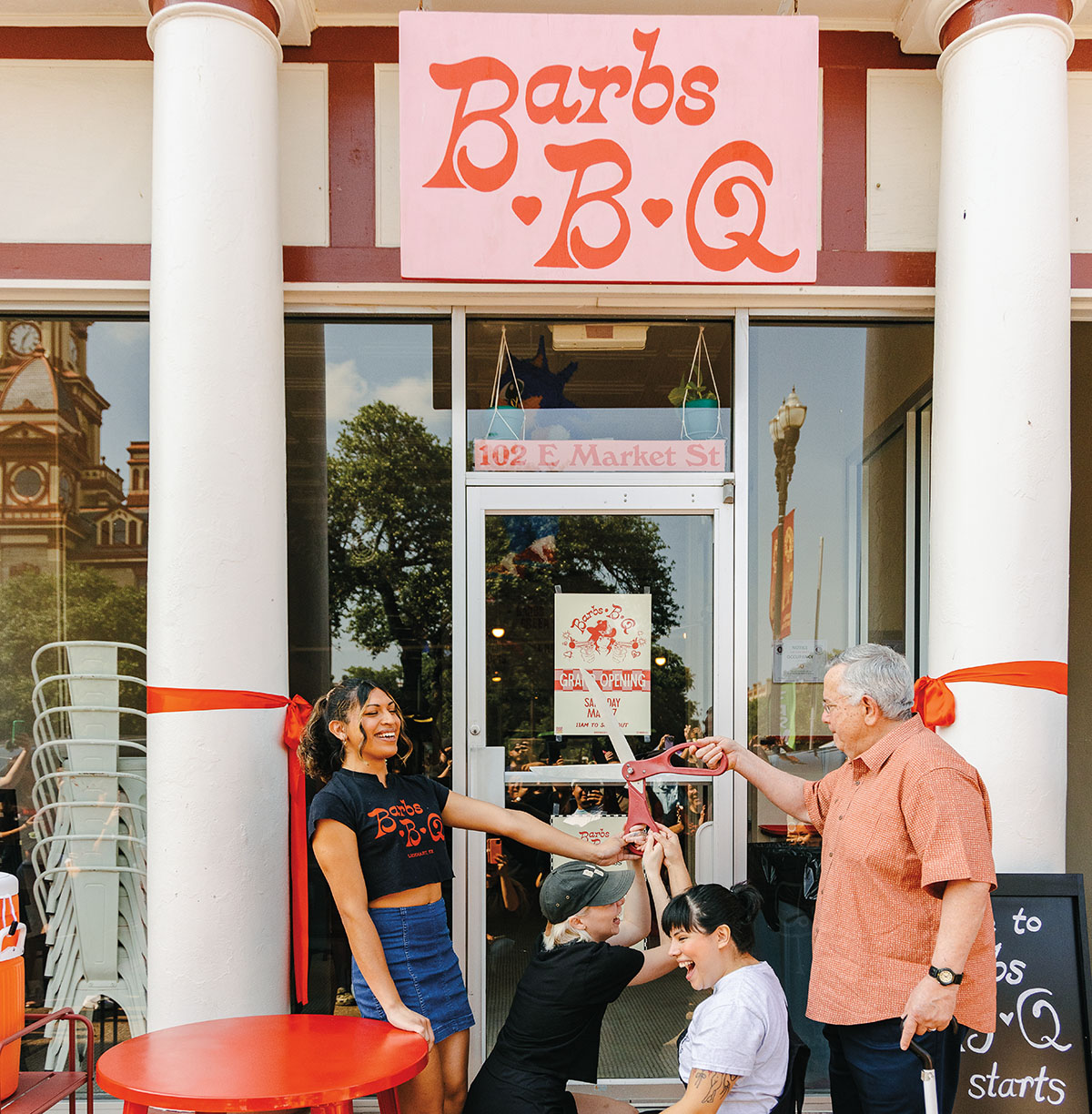 A group of people cut a large ribbon in front of a building with a pink sign reading "Barbs B-Q"