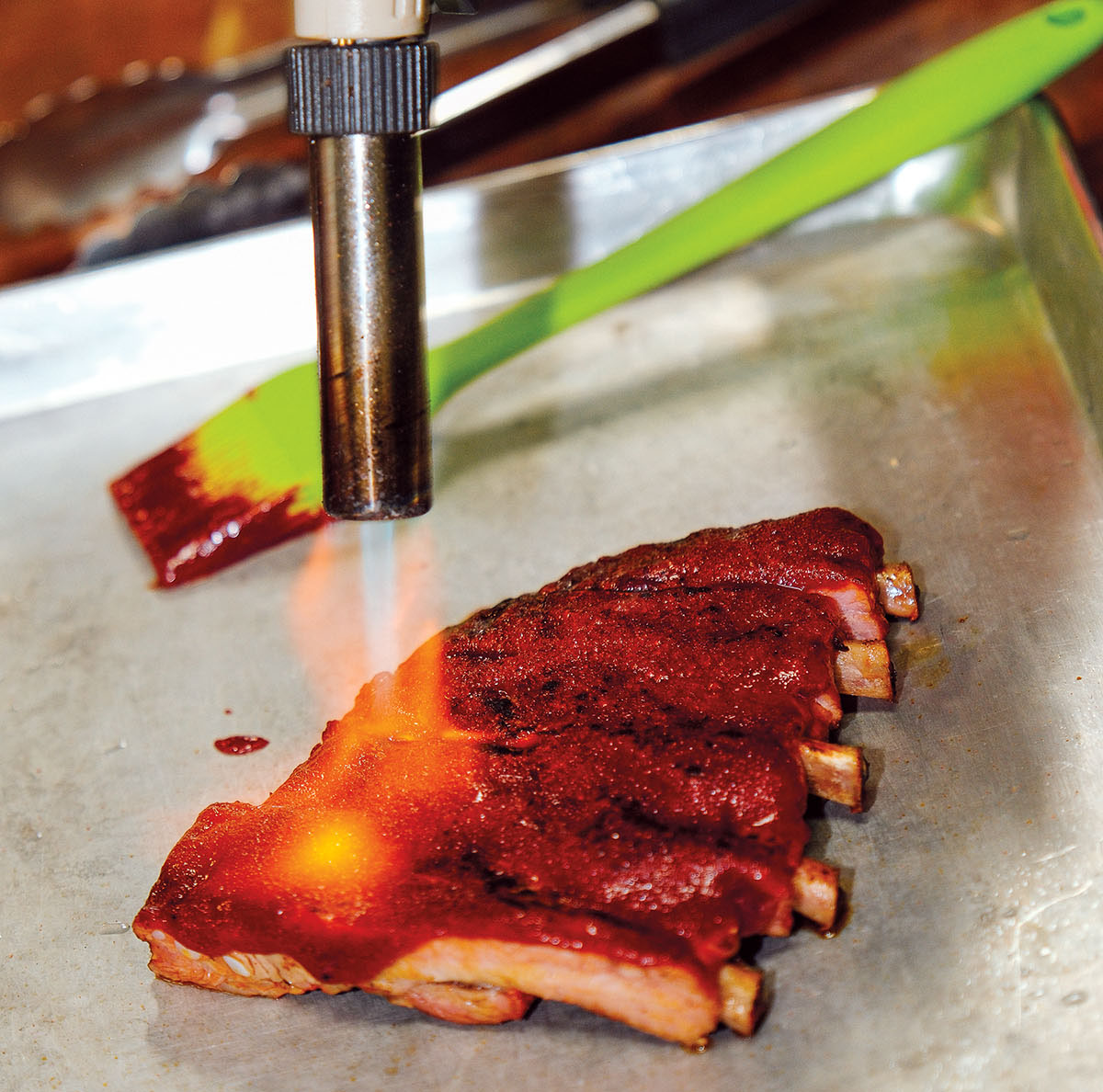 A silver blowtorch is used to apply a flame to a bright red rack of ribs