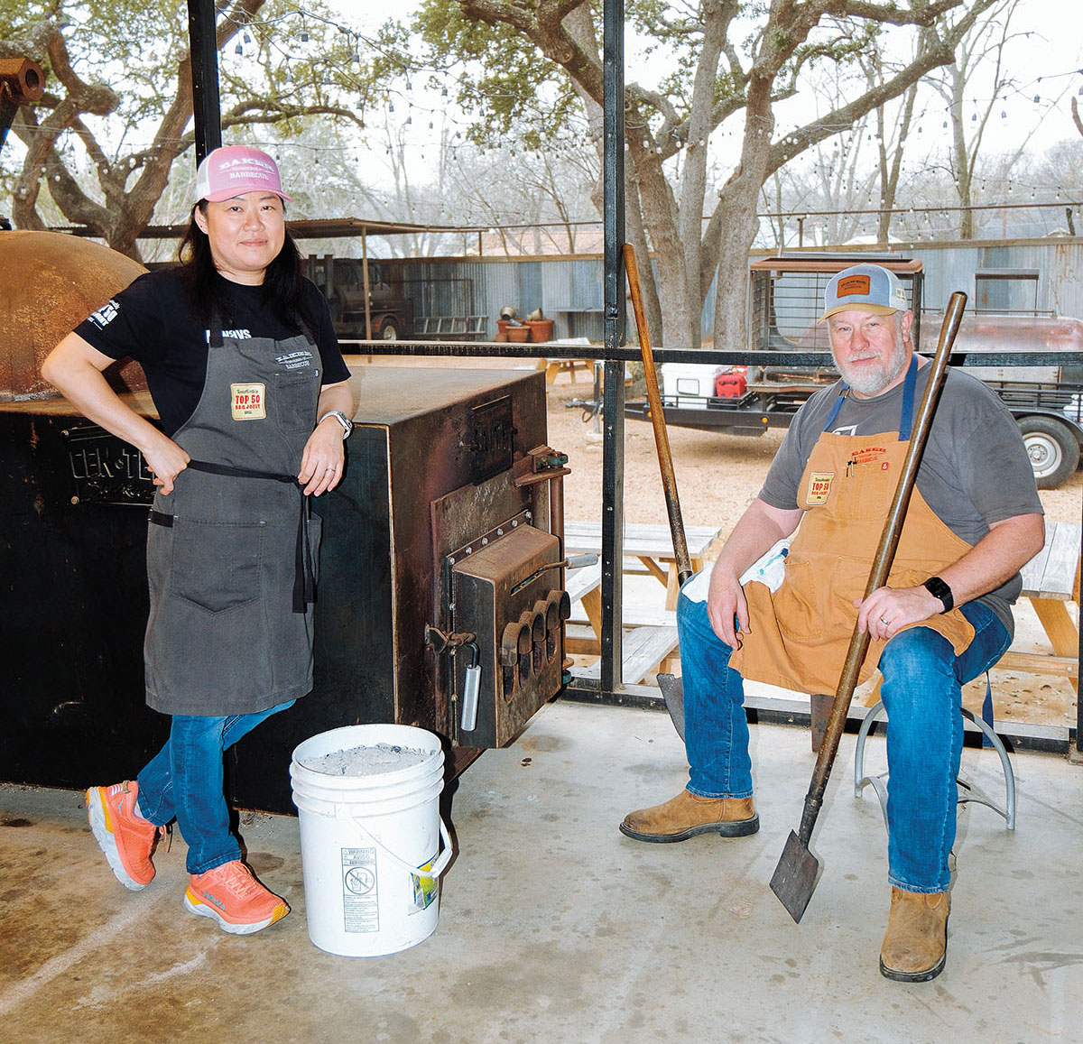 Two people stand next to a barbecue pit smoker