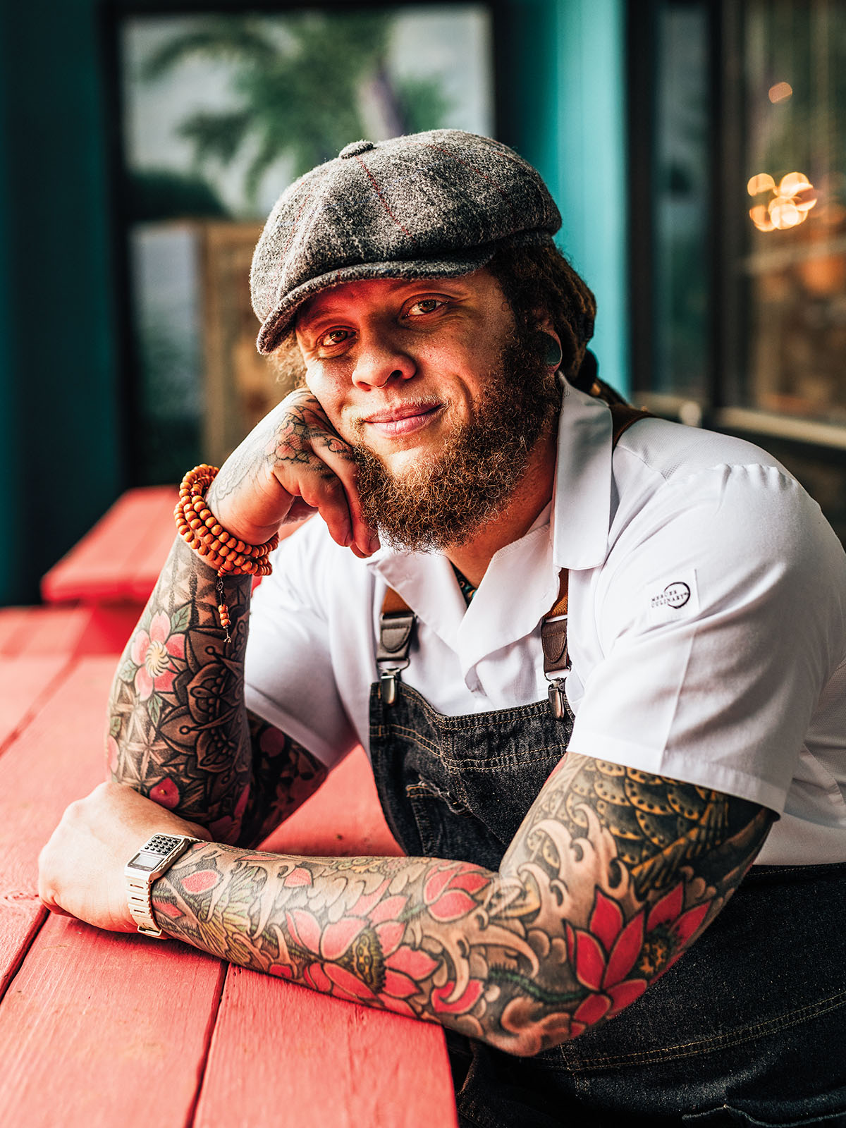 A man with two sleeve tattoos sits at a table in a chef's apron and white shirt