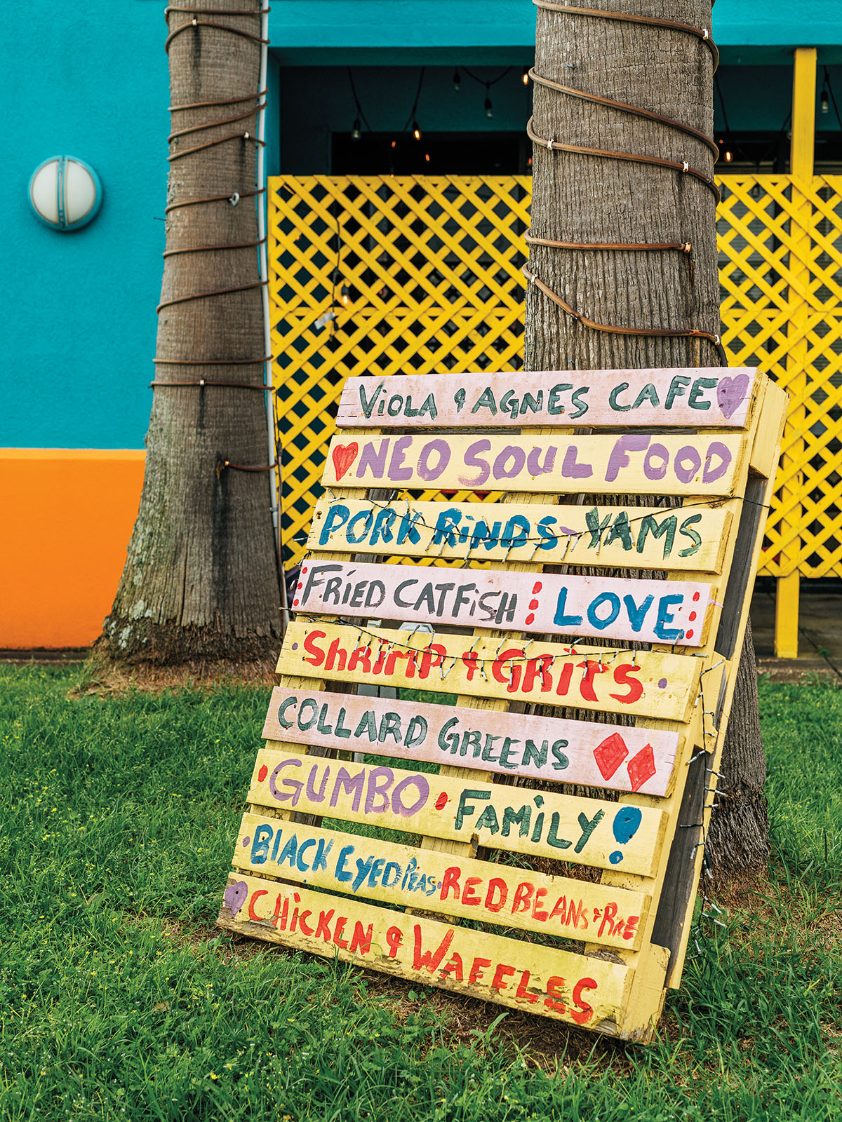 Menu items, including fried catfish, shrimp and grits, collard greens, gumbo, and black eyed peas are hand-painted in bright colors on a wooden pallet