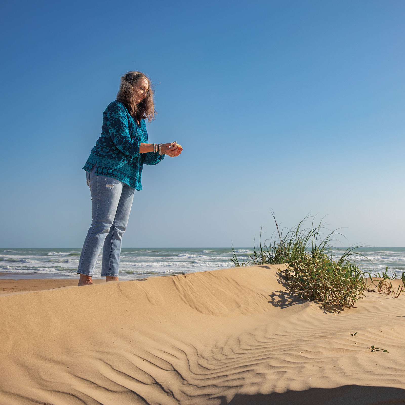 A woman wearing a blue-green flannel shirt stands on a sand dune next to scrub grass