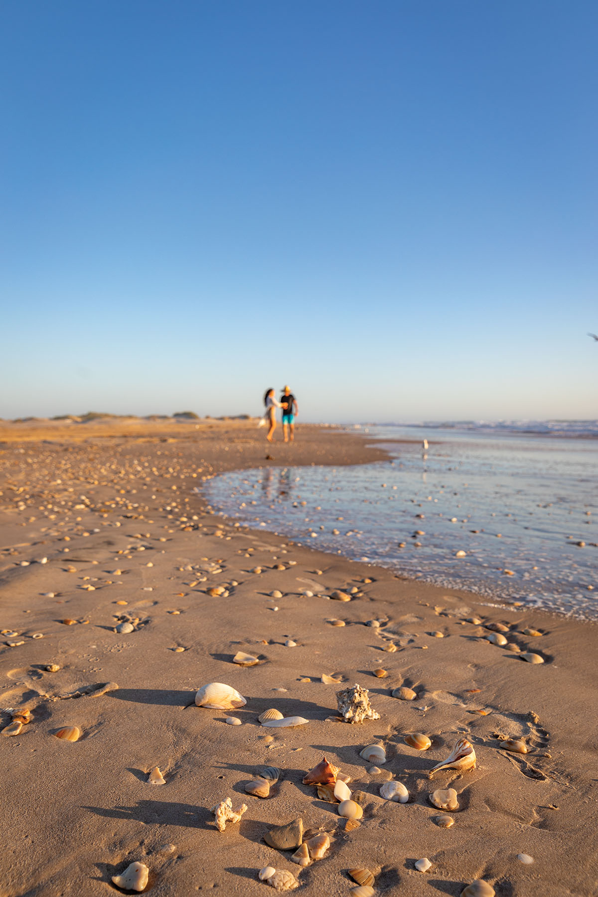 A couple walks along the sand next to clear water under blue sky