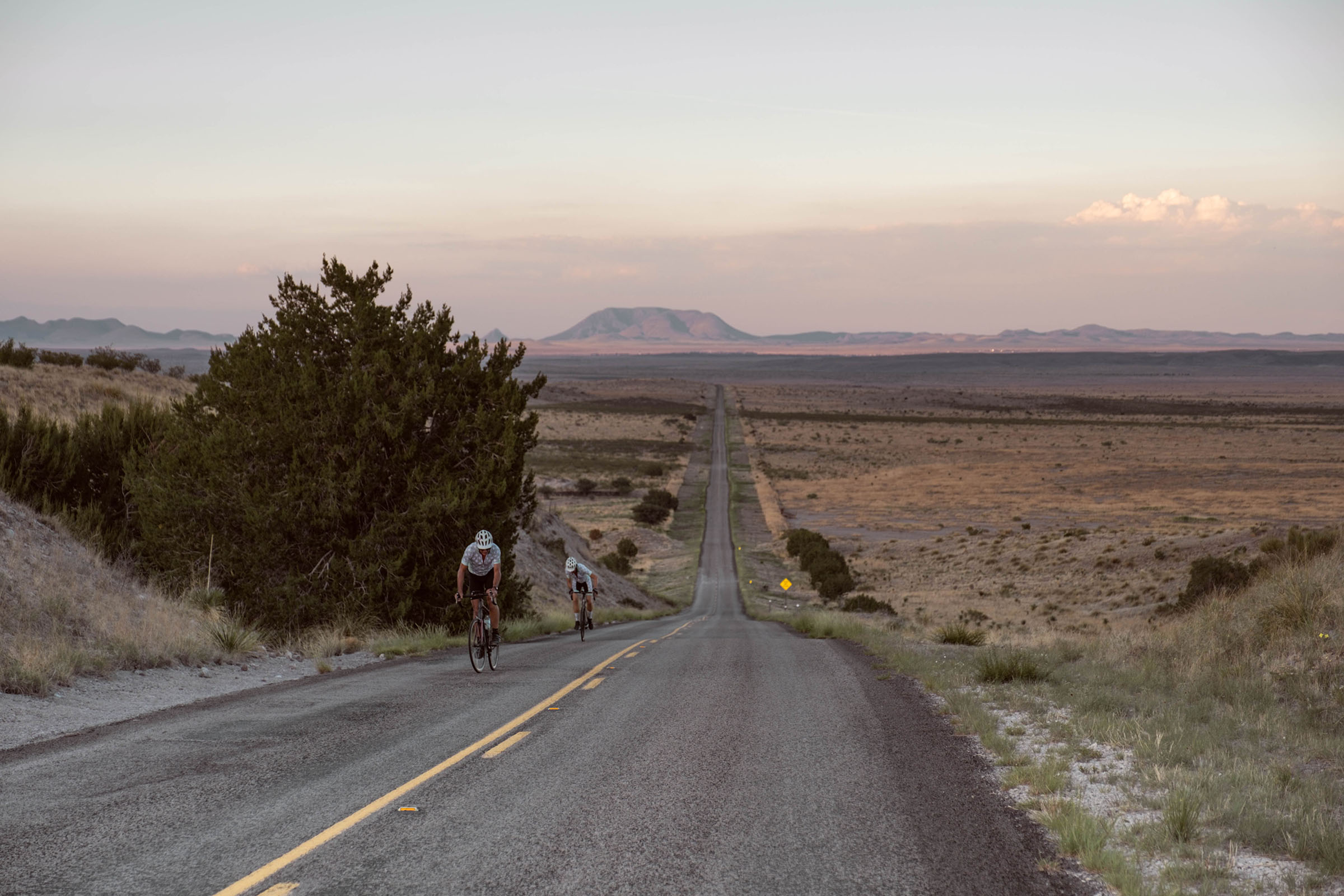 A group of cyclists ride along a desert road with mountains in the background